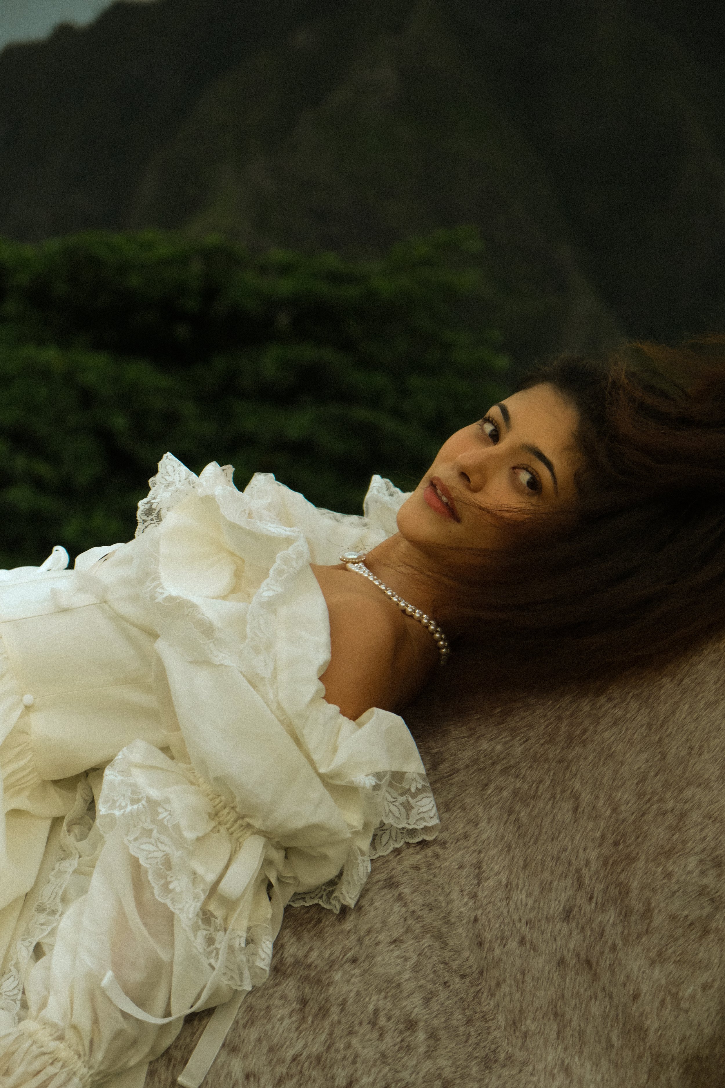 A woman lying on her side on a blanket, wearing a white dress with ruffled sleeves and lace details, with a pearl necklace, dark hair, and a neutral expression, outdoors in front of a mountain landscape.