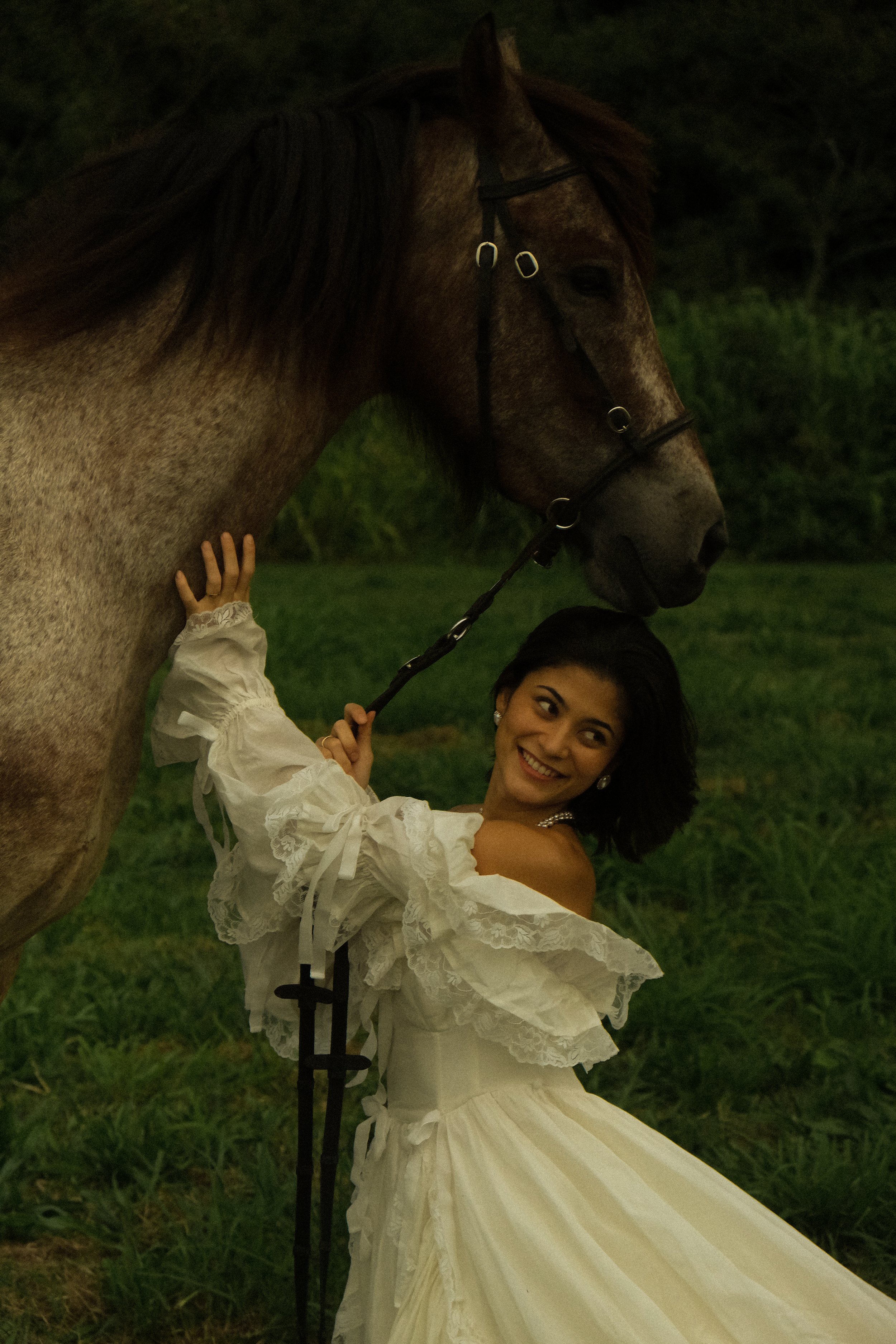 A woman with dark hair and earrings wearing a white dress with ruffled sleeves. She is smiling and holding a lead rope of a brown horse with a black mane, which is close to her face. The background shows green grass and trees, indicating an outdoor s