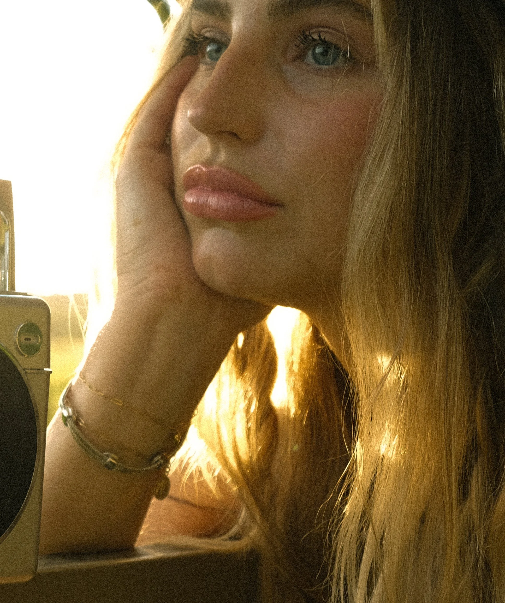 A close-up of a young woman with long blonde hair, blue eyes, and light skin, resting her face on her hand, looking contemplative.