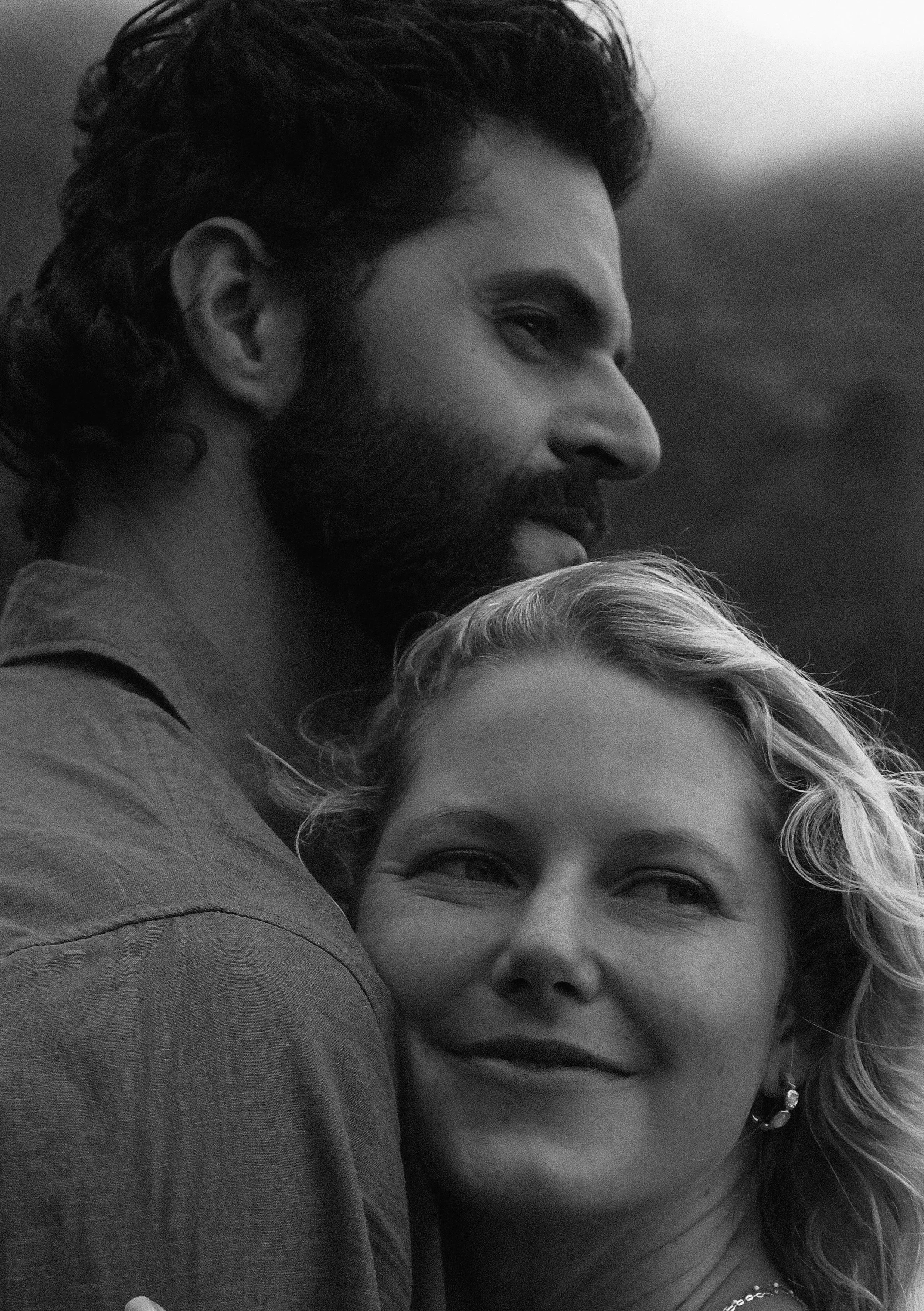 Close-up of two people, a man with dark, curly hair and a beard, and a woman with light-colored hair and earrings, embracing closely.
