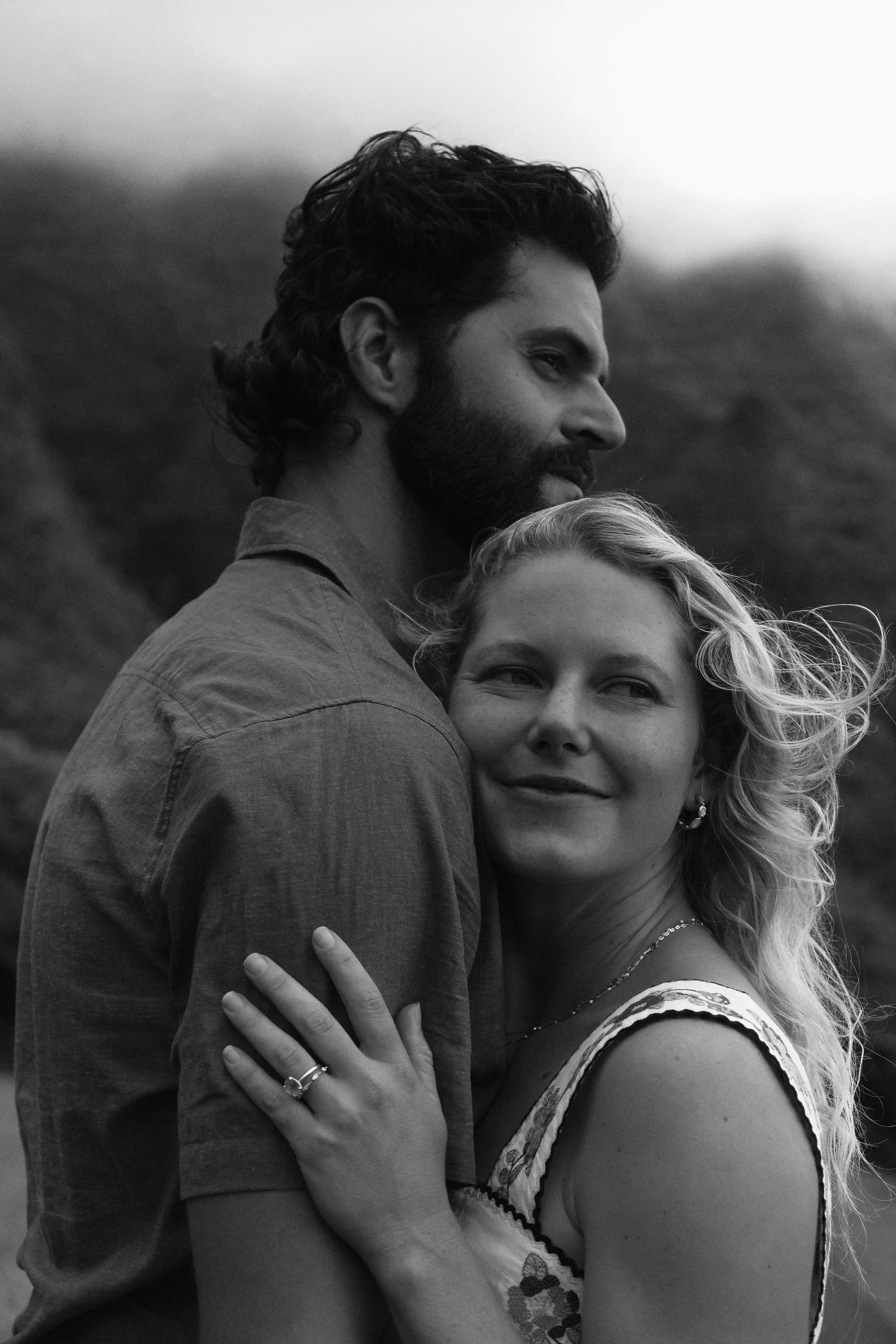 Black and white photo of a man and woman embracing each other outdoors. The man has dark, wavy hair and a beard, and the woman has blonde, curly hair. They are smiling and looking off into the distance.