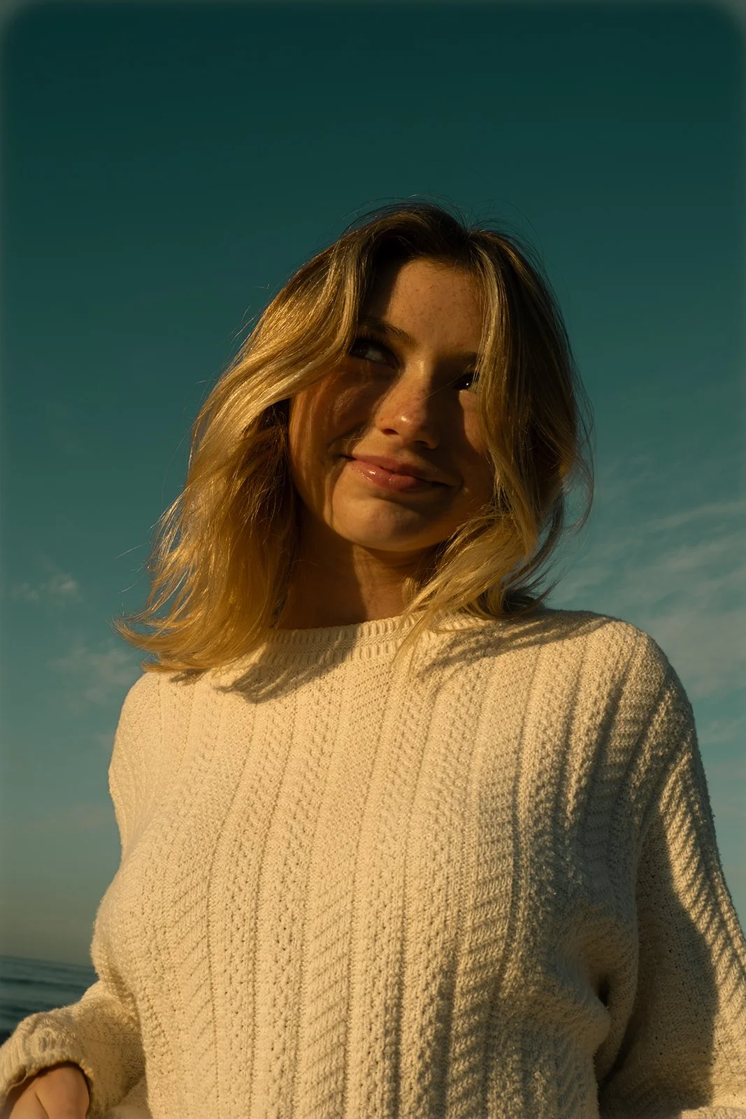 A woman with blonde hair wearing a cream-colored sweater, smiling, outdoors with a blue sky background.