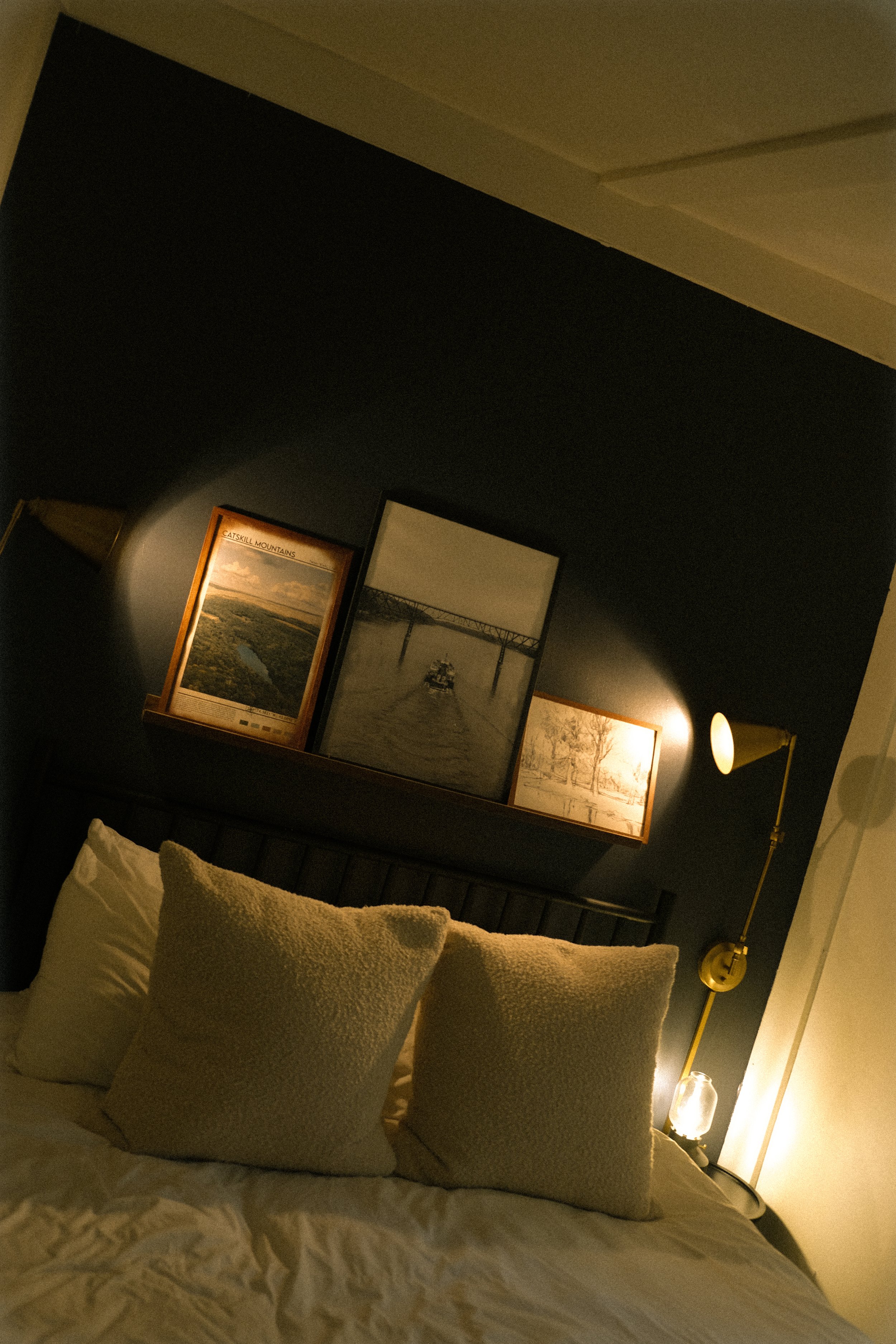 Close-up of a bed with pillows in a dimly lit room. On the dark wall behind, there are framed pictures and a wall-mounted lamp and a floor lamp providing warm lighting.