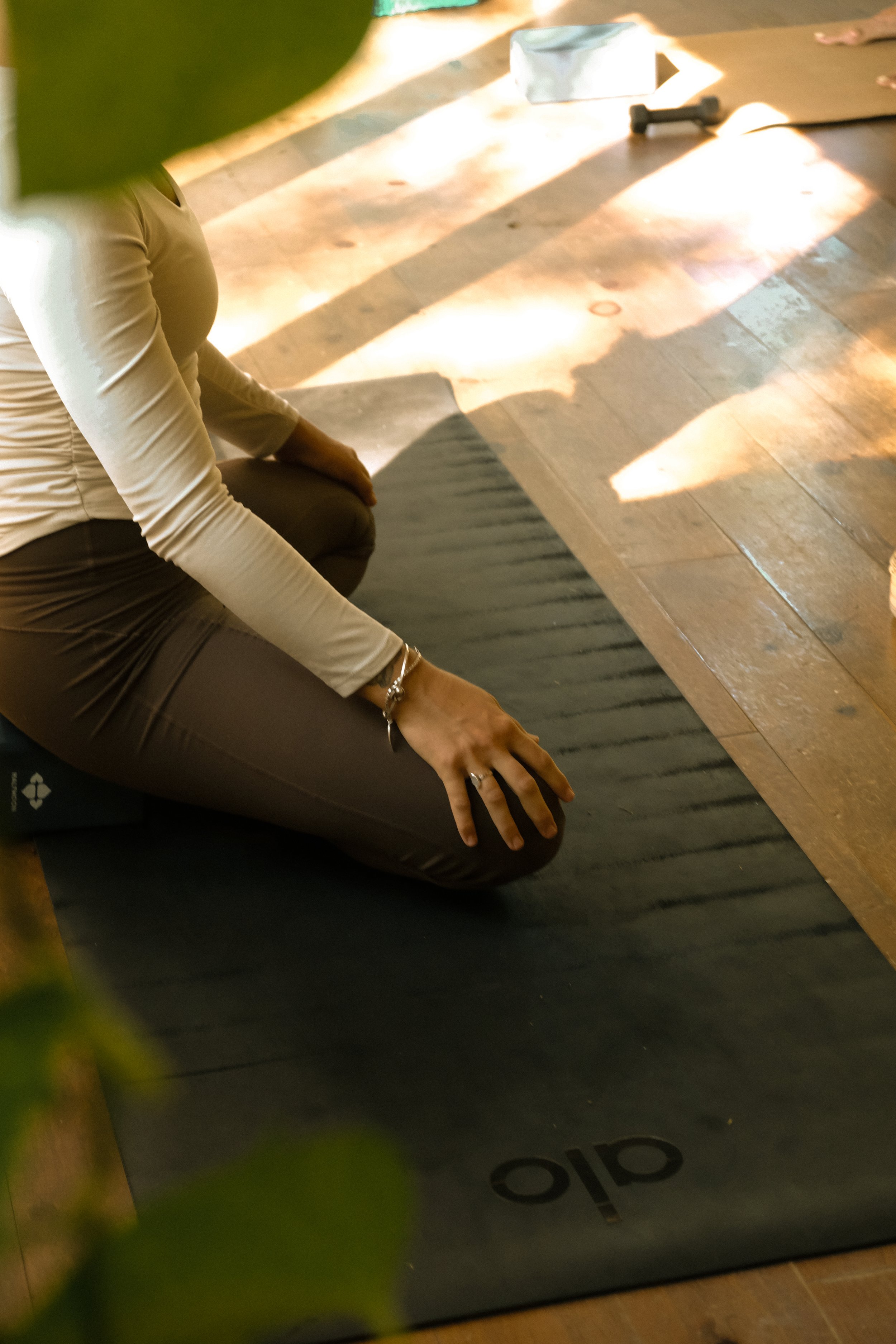 Person sitting cross-legged on yoga mat in a sunlight-filled room, with part of a yoga block, roller, and some exercise equipment visible in the background.