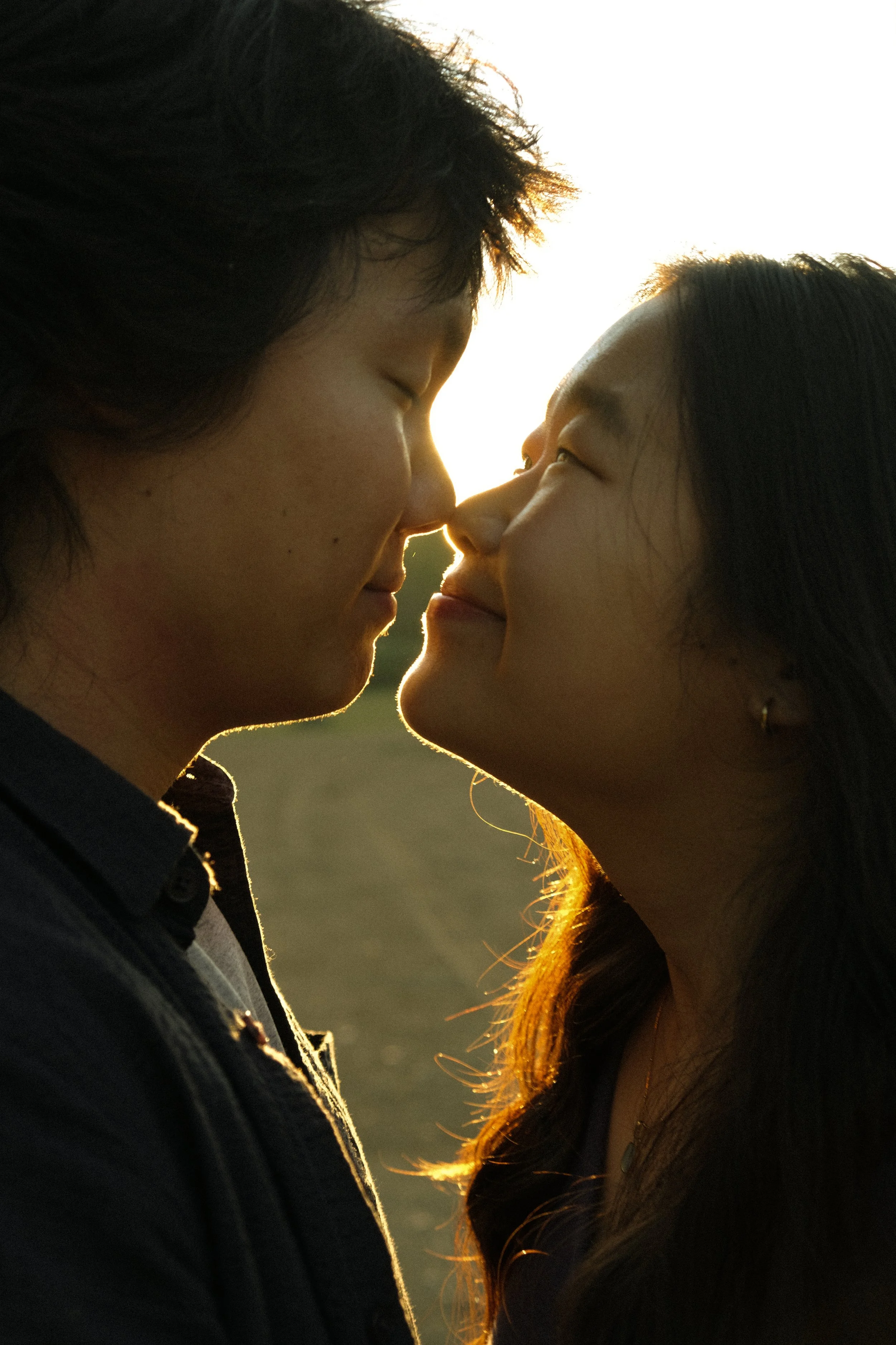 Close-up of two people touching noses, smiling, outdoors at sunset, backlit with warm sunlight.