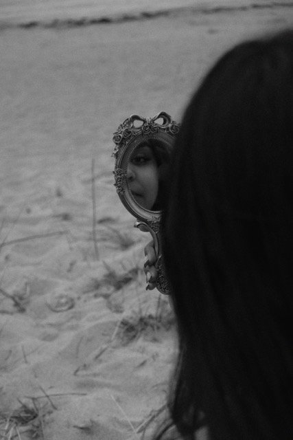 Person with long hair looking into an ornate hand mirror on sand, capturing part of their face in the reflection.