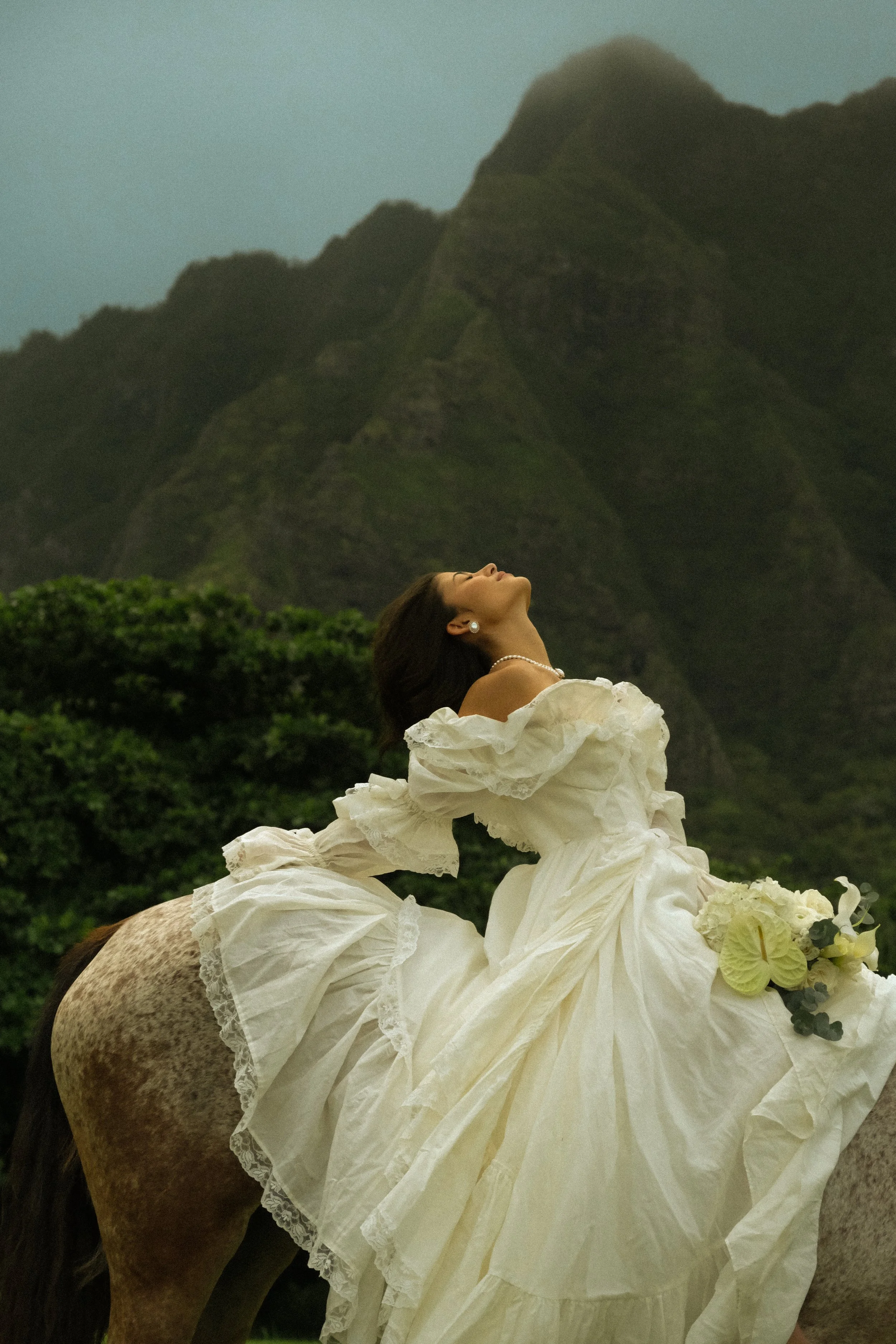 Close-up of a woman with dark hair and earrings, wearing a white dress, with her head tilted back against a mountain landscape background.