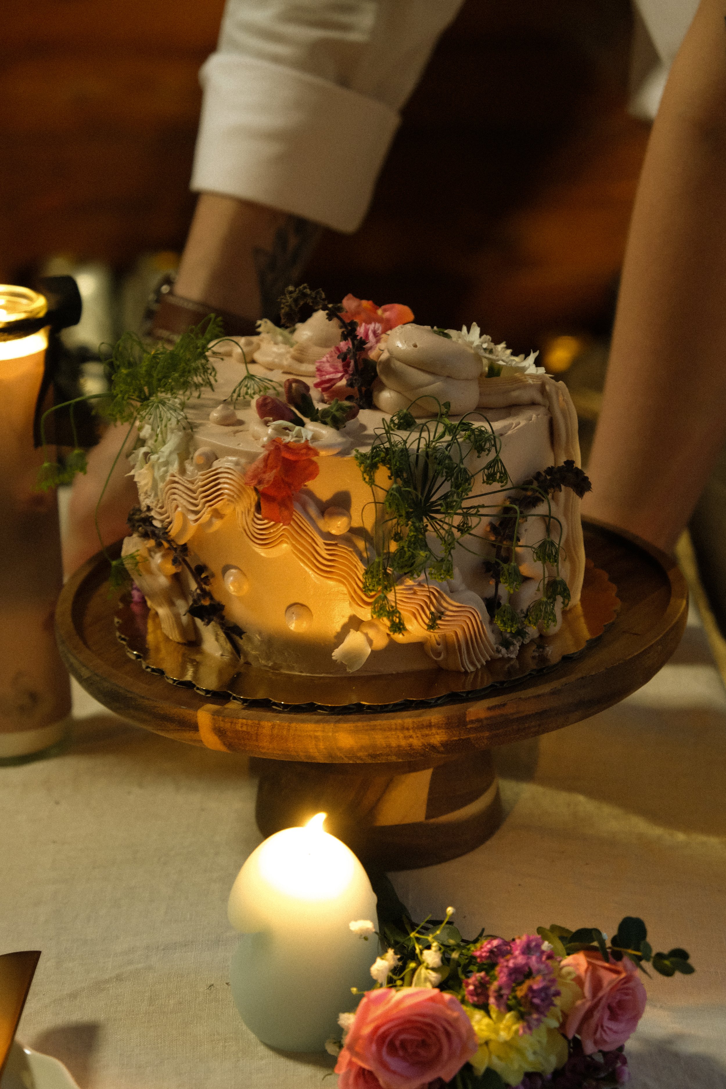 A decorated cake on a wooden stand surrounded by pink and white flowers with a lit white candle in front.
