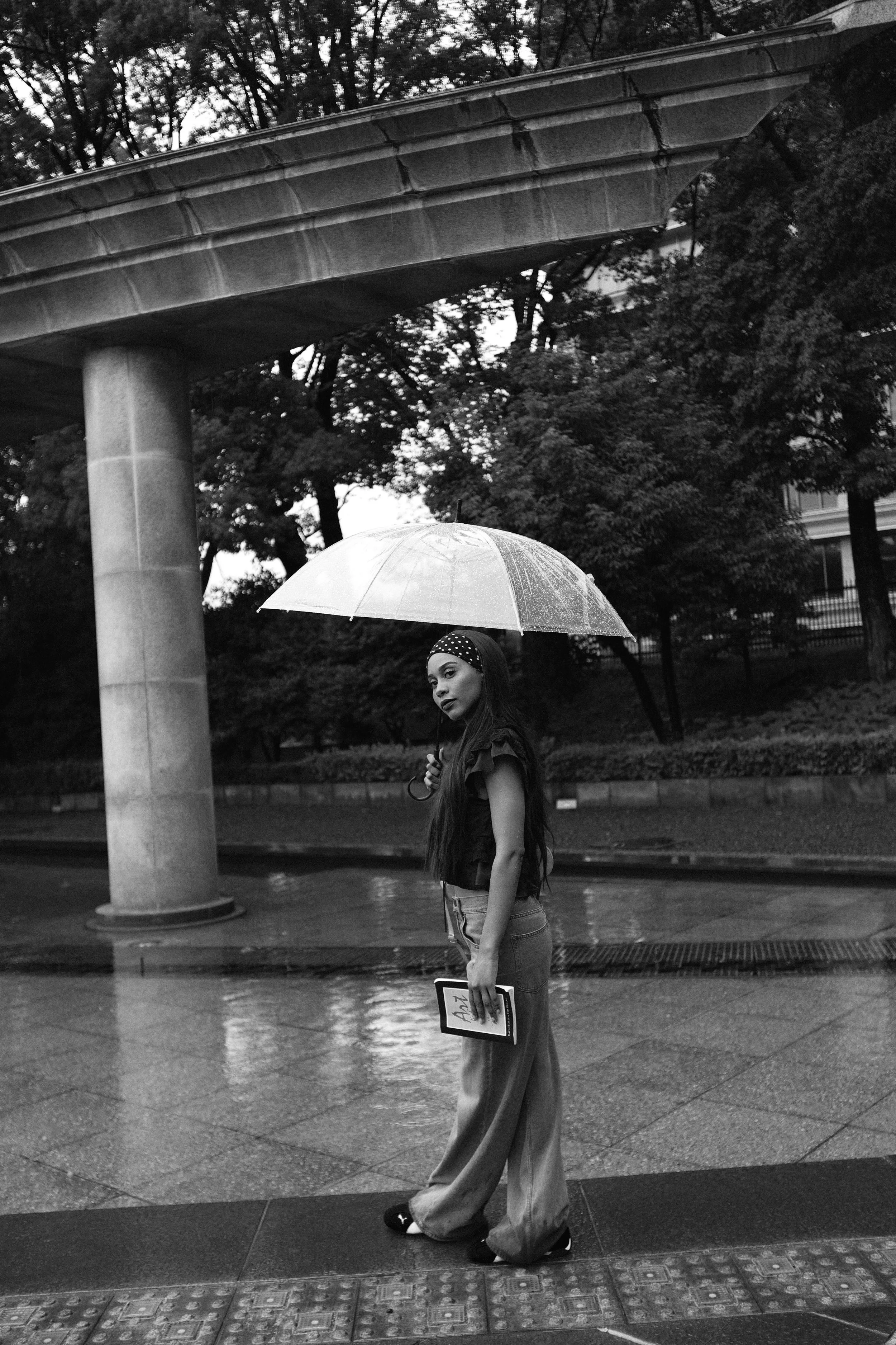 A woman standing on a rainy city street holding a clear umbrella and a book, with a large concrete structure and trees in the background.