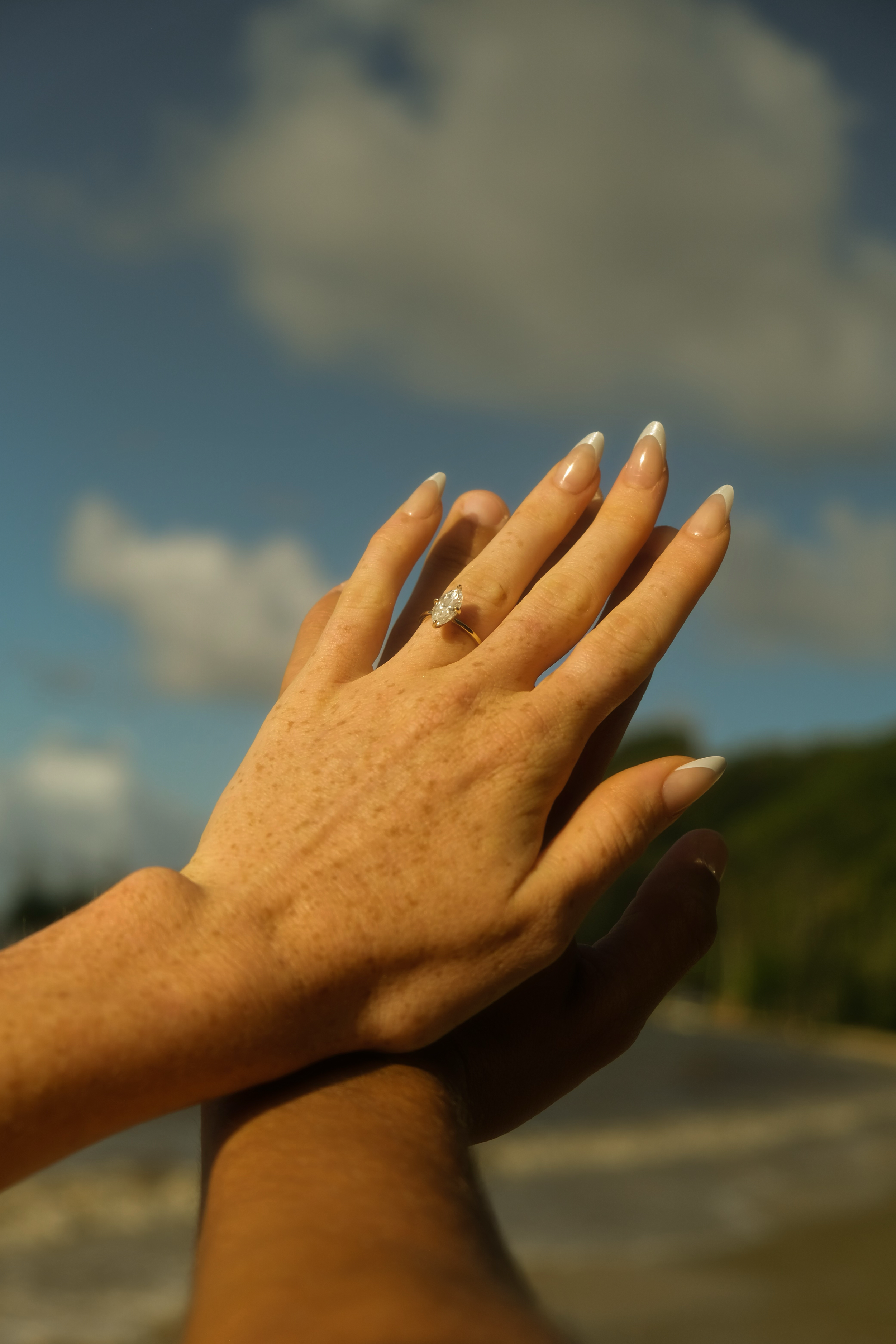 A person's hand with a ring is held up against a cloudy blue sky, with the other hand below supporting it, possibly during the daytime.
