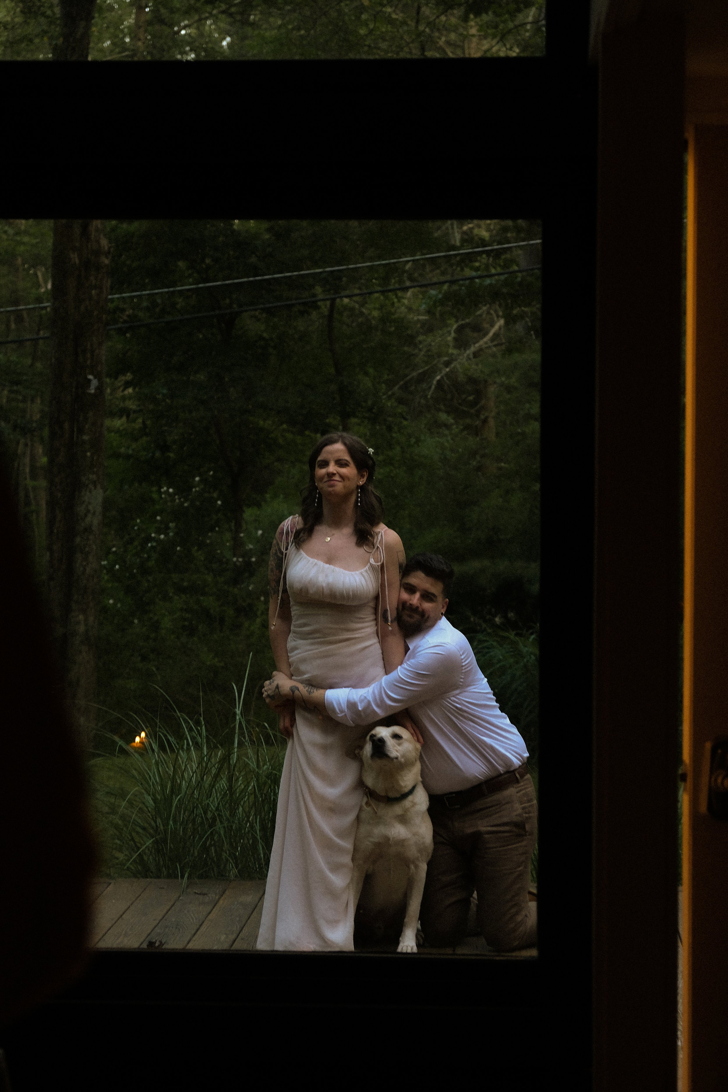 A woman in a white dress, a man in a white shirt and khaki pants, and a Labrador Retriever dog stand outside among trees, with the image framed by a window or door.