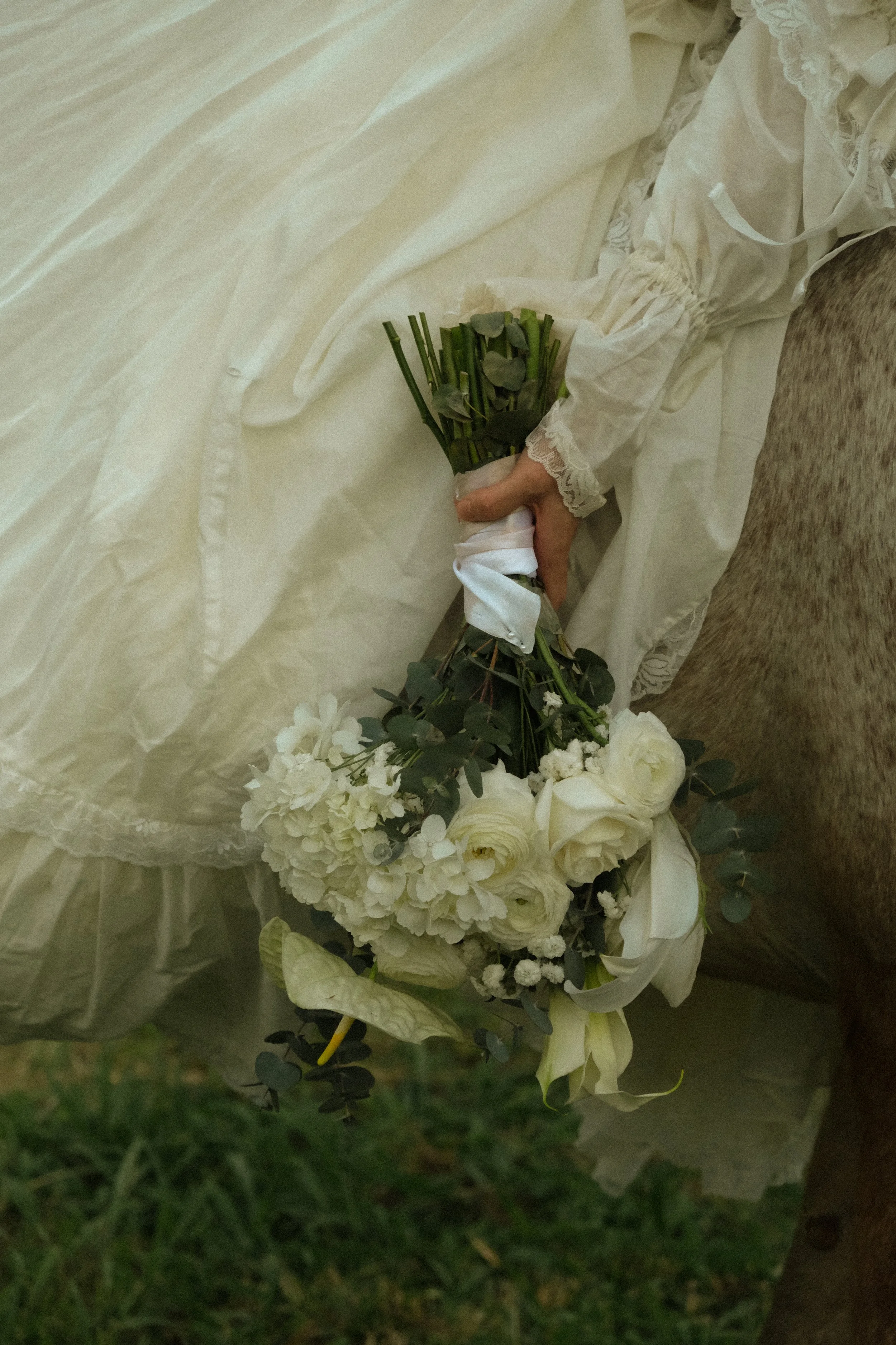 Person holding a bouquet of white and green flowers, including roses and hydrangeas, wrapped in white paper, with the person wearing lace-trimmed white clothing.