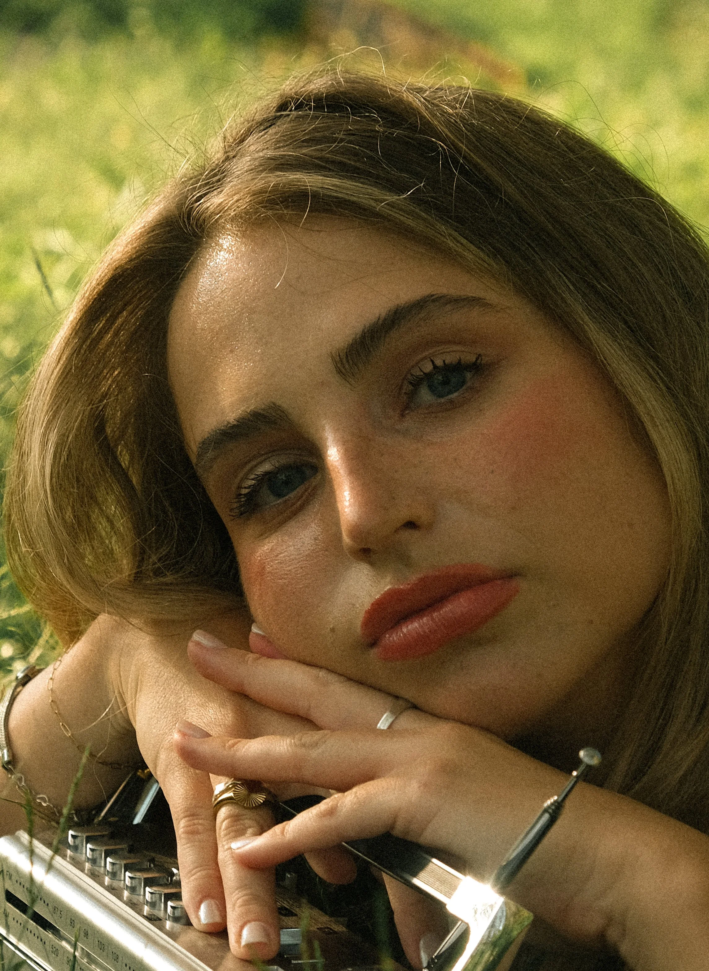 Close-up of a woman's face with blue eyes and blonde hair, resting her hand on her face while lying on a typewriter keyboard, with a blurred green background.