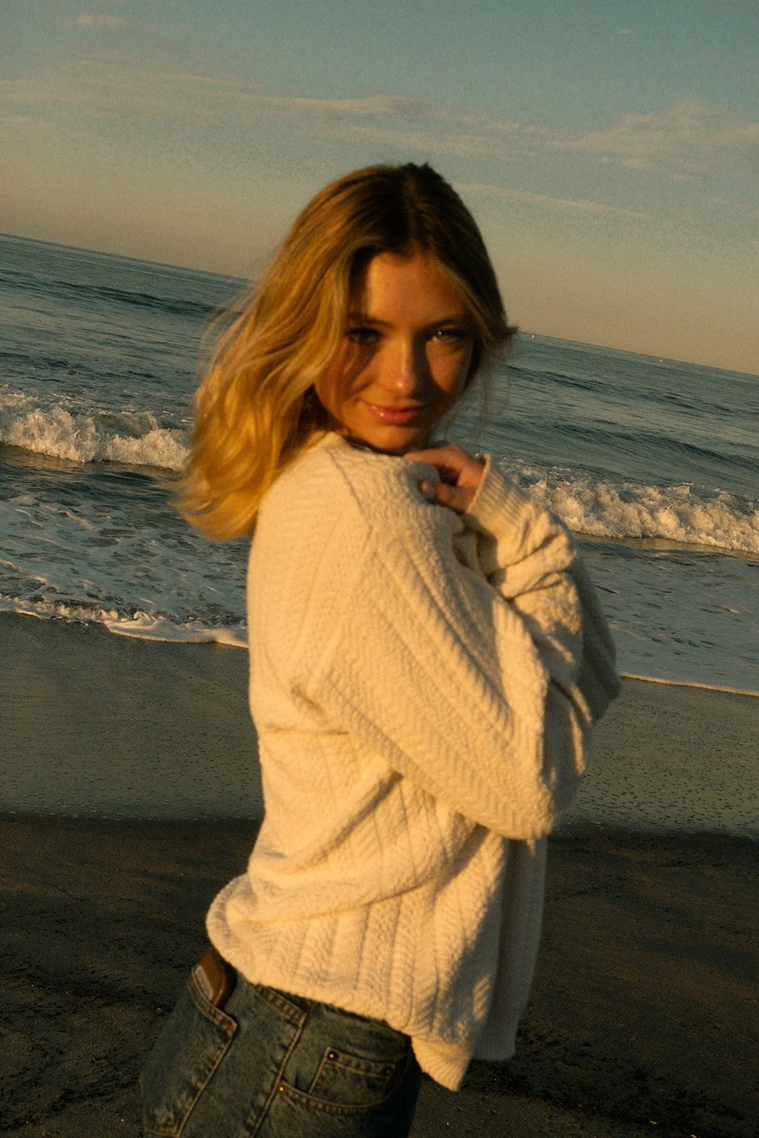 A young woman with shoulder-length blonde hair standing on a beach during sunset, wearing a cream-colored sweater and jeans, with ocean waves in the background.
