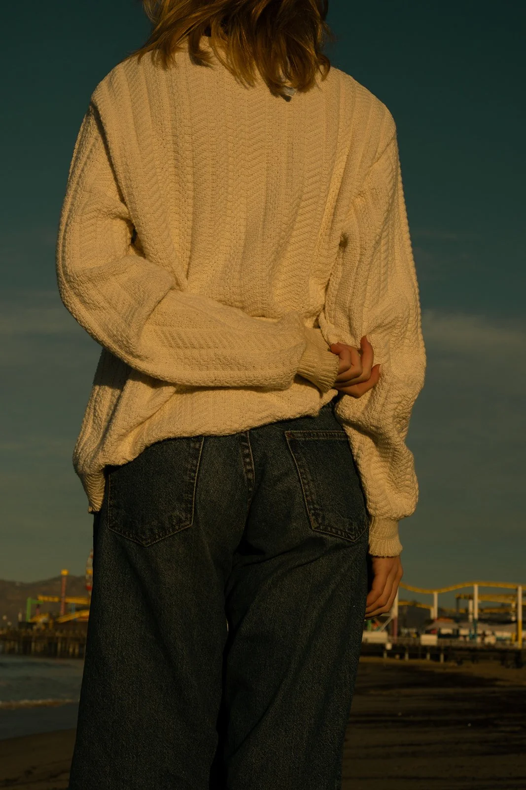 A woman wearing a beige sweater and black jeans stands near the beach during sunset, with a pier and amusement park structures in the background.