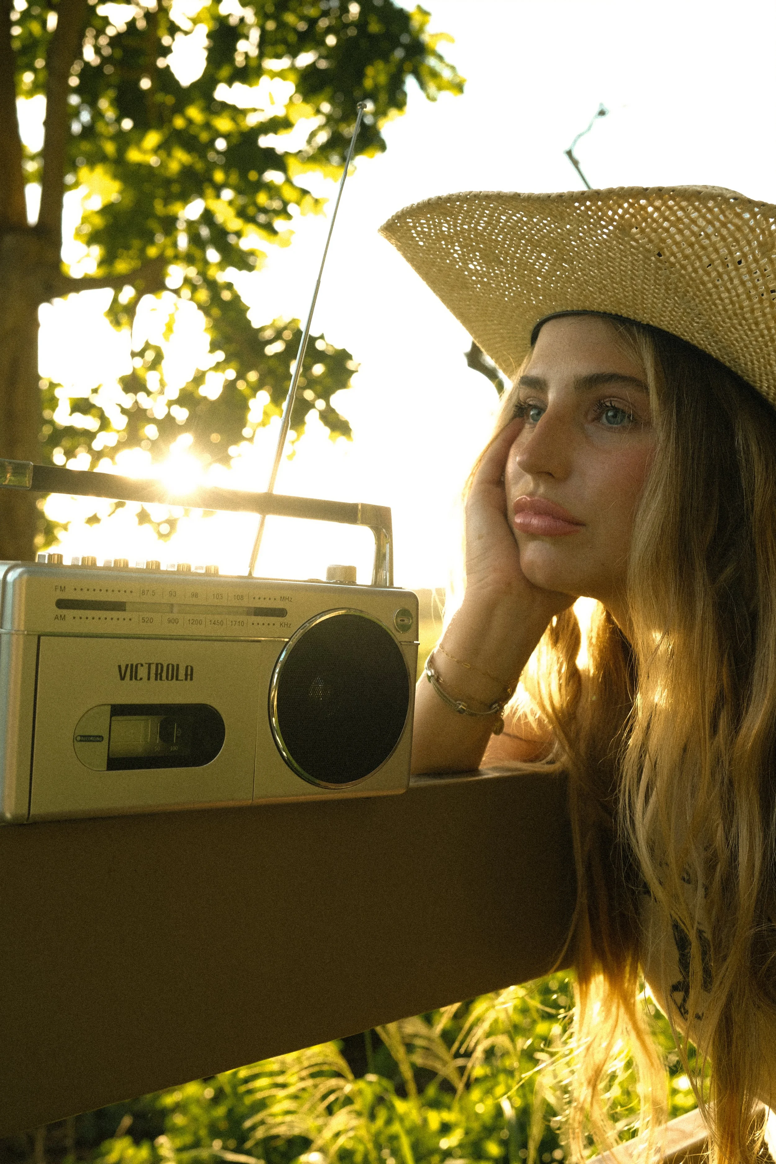 A young woman with long blonde hair and blue eyes wearing a large straw hat and resting her head on her hand. She is outdoors with sunlight filtering through the trees in the background. In front of her, there is a vintage-looking radio.