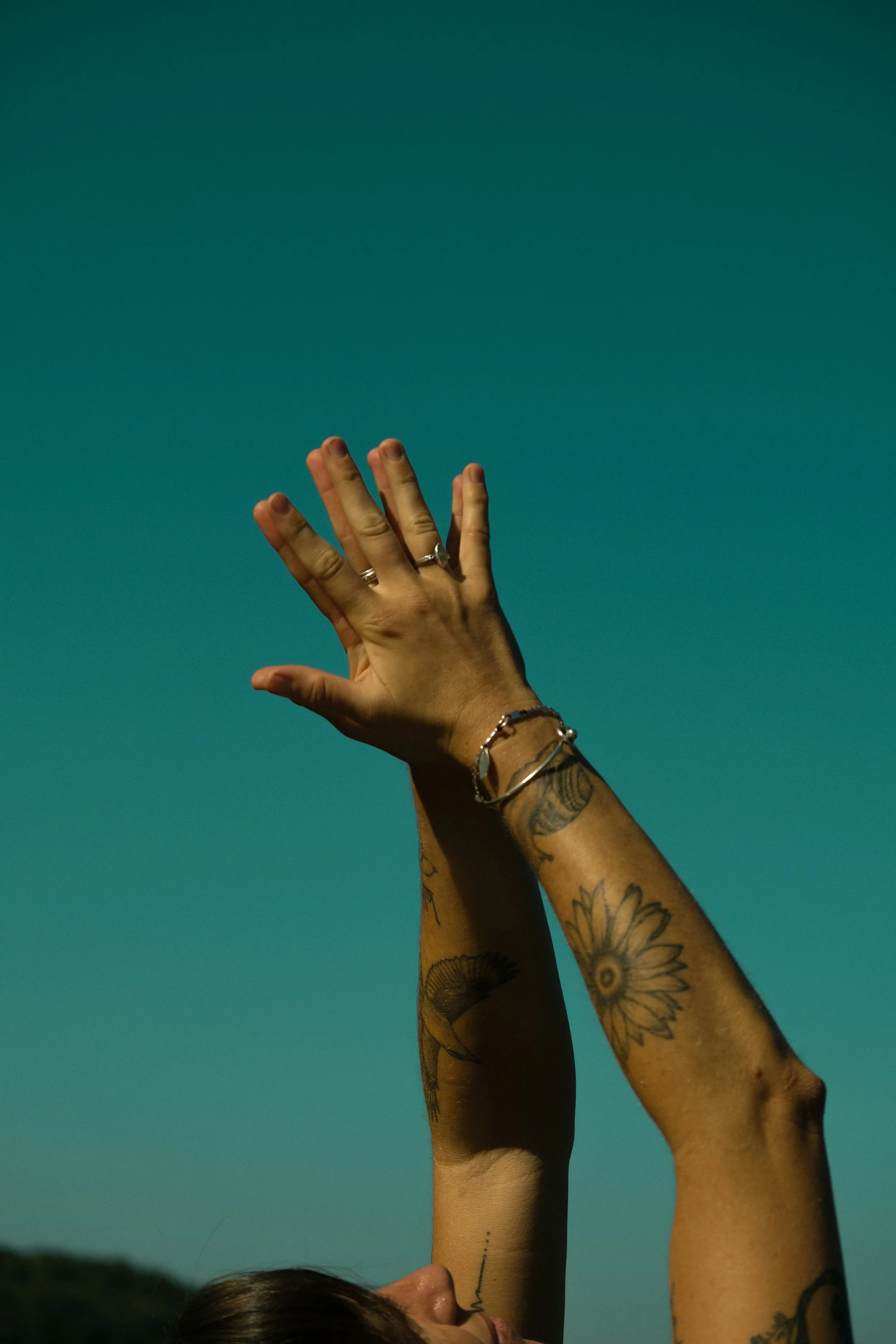 A person with tattoos on their arms and a silver bracelet is extending their hand and arm upward against a teal sky, with part of their face visible at the bottom of the image.