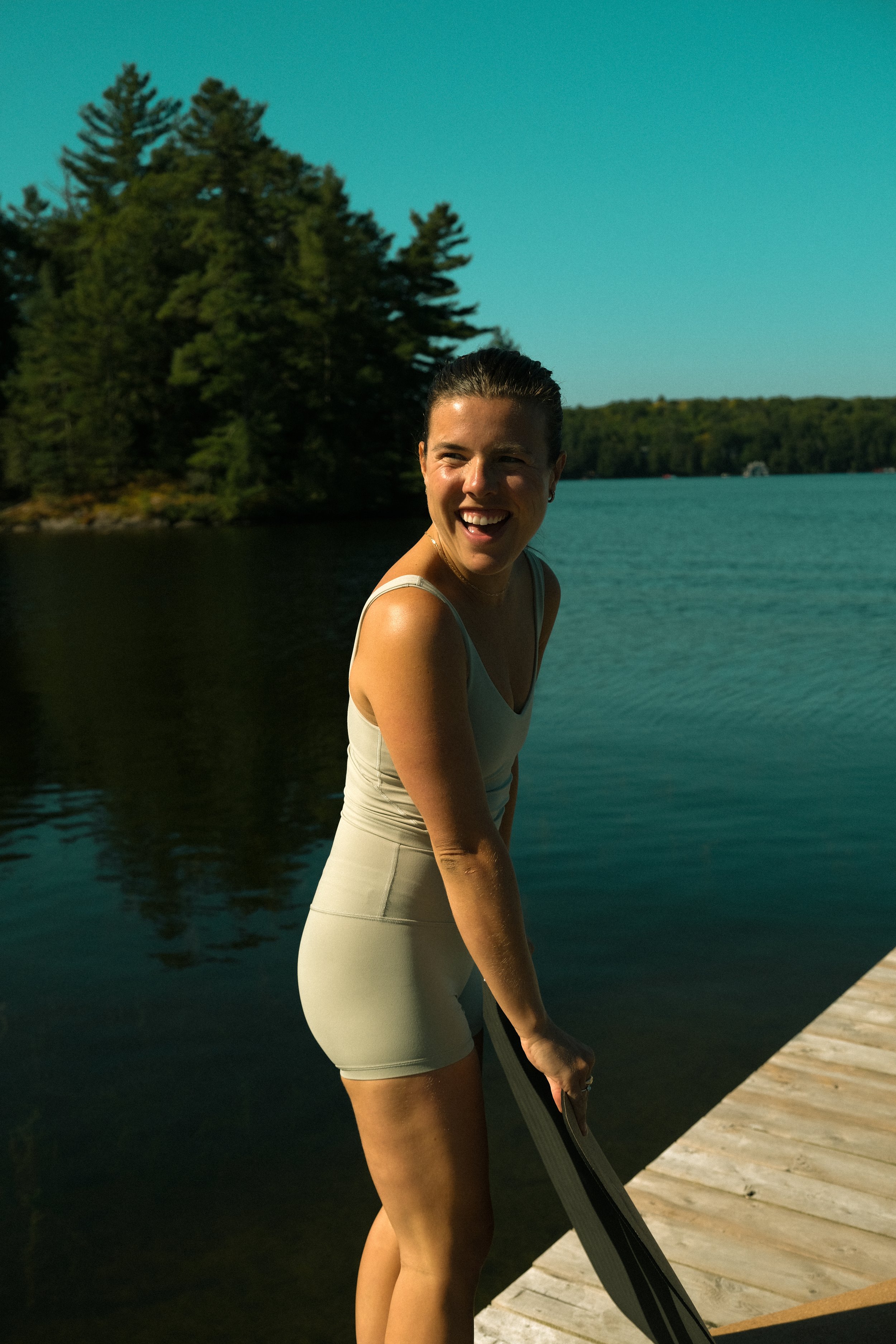 A woman in a beige dress standing on a dock by a lake, smiling, with trees in the background under a clear blue sky.