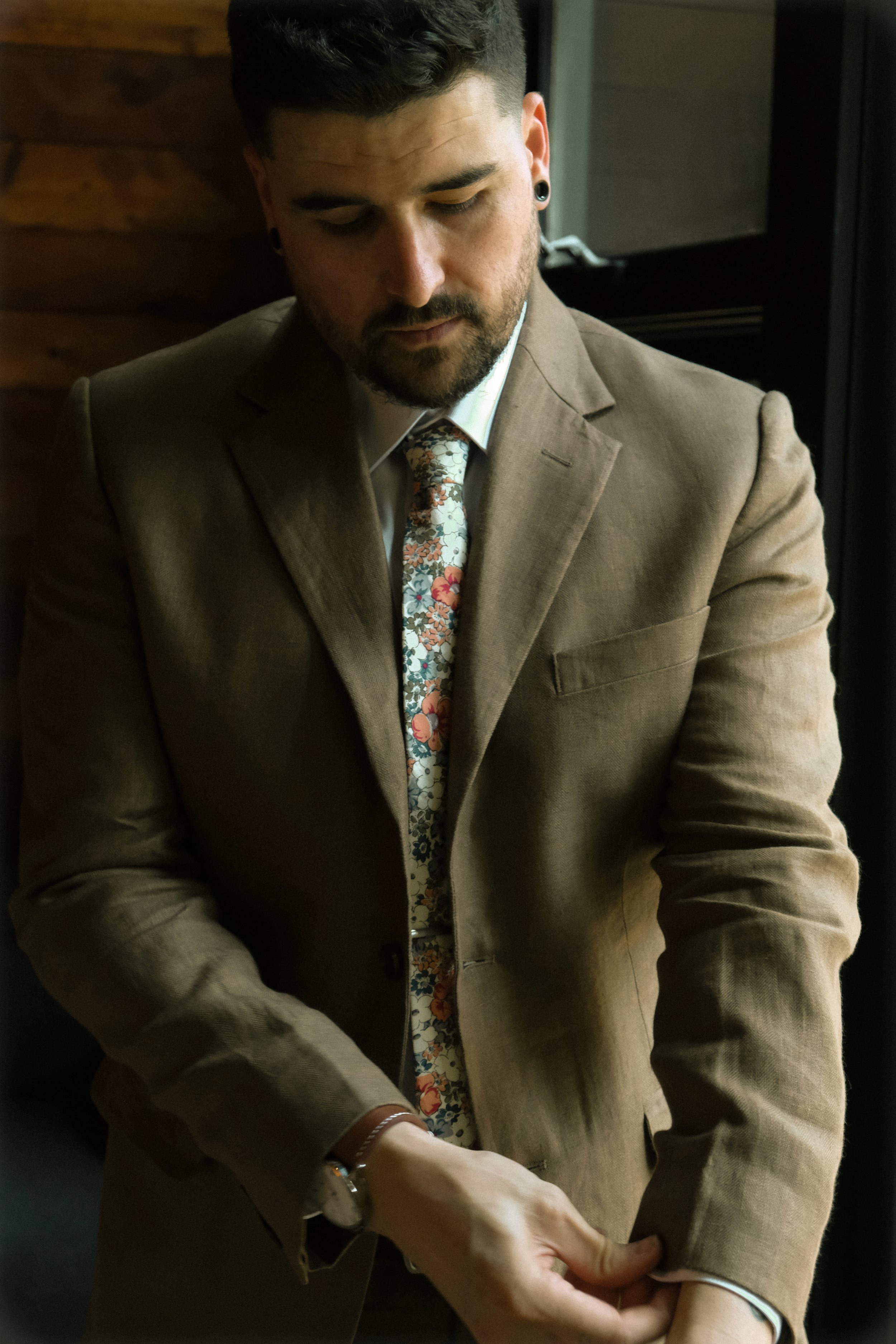 A man with dark hair, beard, and earrings wearing a tan blazer and floral tie, appears to be sitting with his head slightly bowed, with a woman’s hand holding his wrist, in a warmly lit indoor setting.