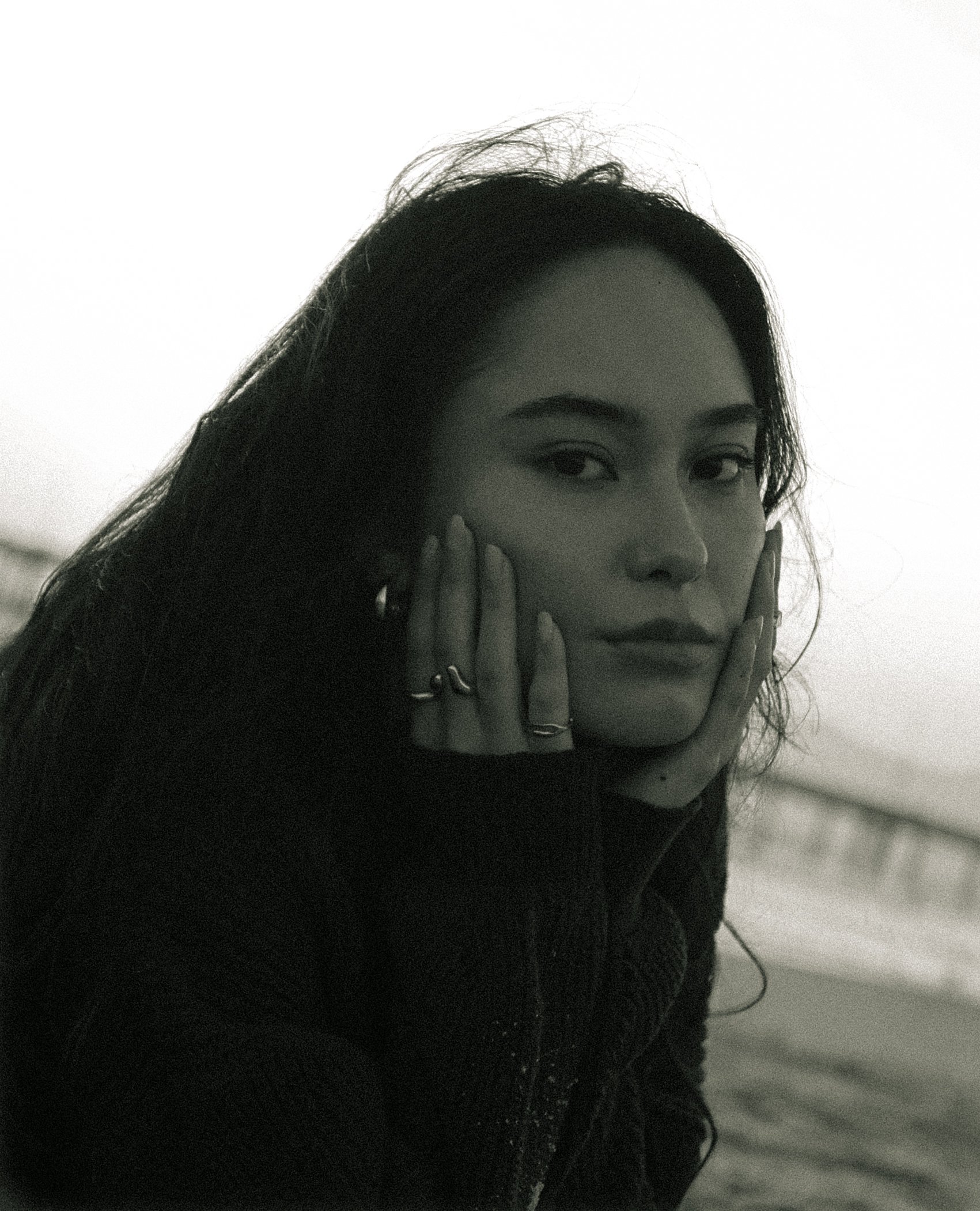 Black and white close-up photo of a woman with dark hair, resting her face on her hands, with visible eyebrows and eyes.