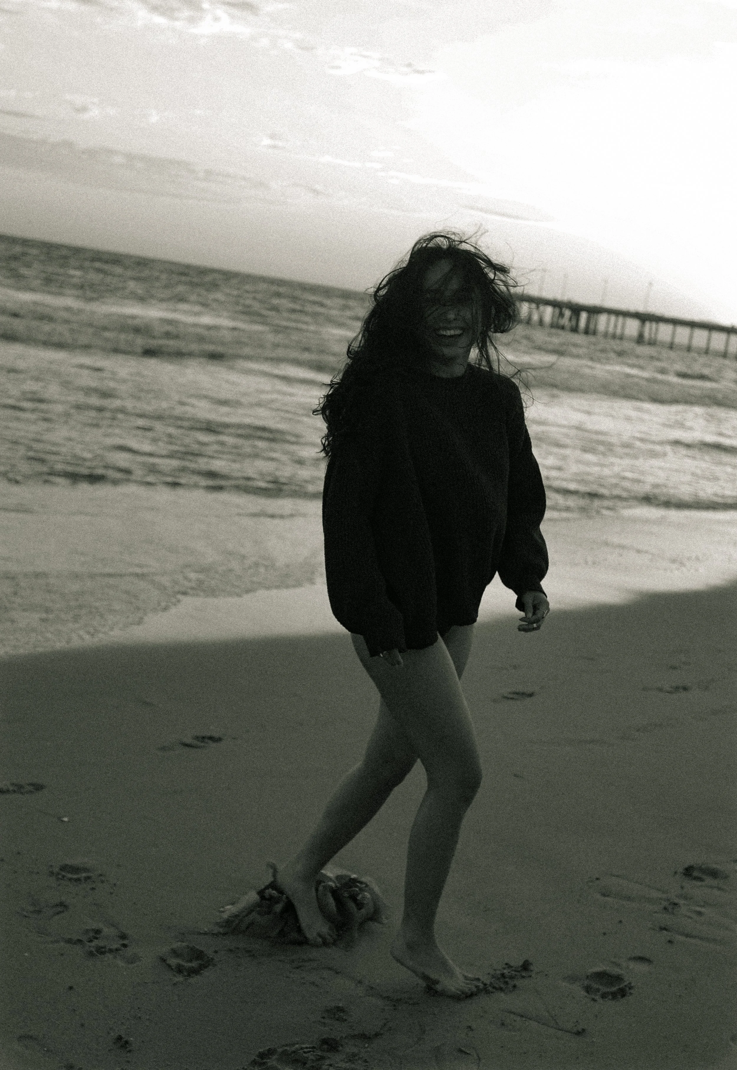 A person with long curly hair standing on the beach with an ocean and pier in the background. The person appears to be smiling and is dressed in dark clothing, with one leg bent and partially in motion, creating a playful and carefree scene.