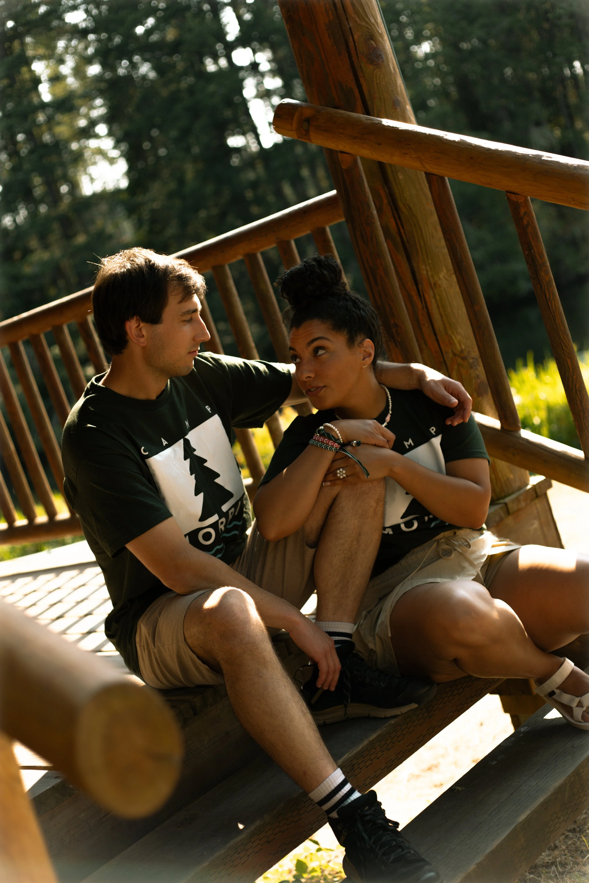 A young man and woman sitting close on a wooden bench outdoors, with the man holding the woman's leg, both wearing black T-shirts with a tree logo and text, surrounded by green trees.