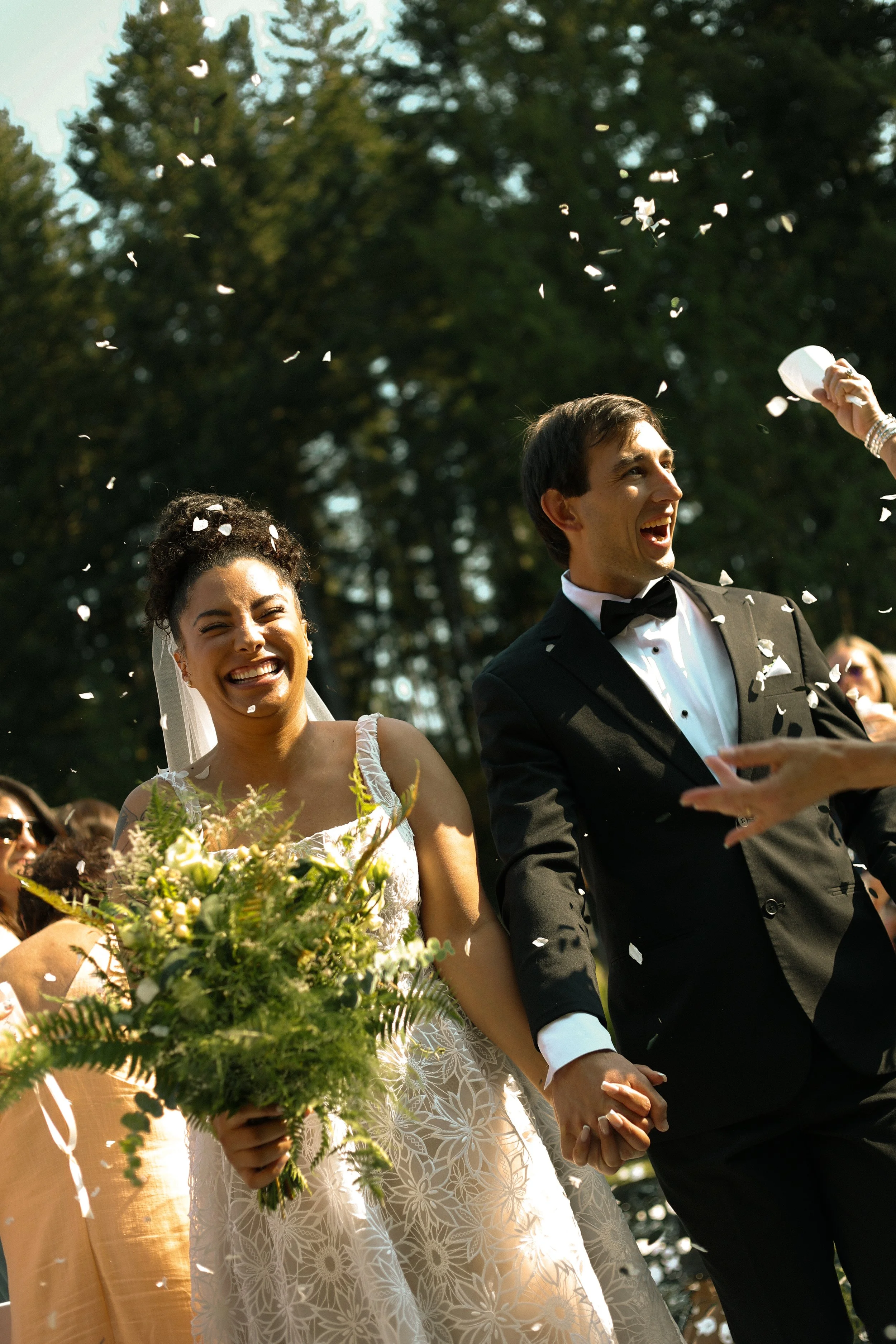 A smiling bride and groom holding hands, outdoors on a sunny day, with white flower petals falling around them, surrounded by wedding guests. The bride wears a white dress with floral pattern and holds a bouquet of green flowers. The groom wears a bl