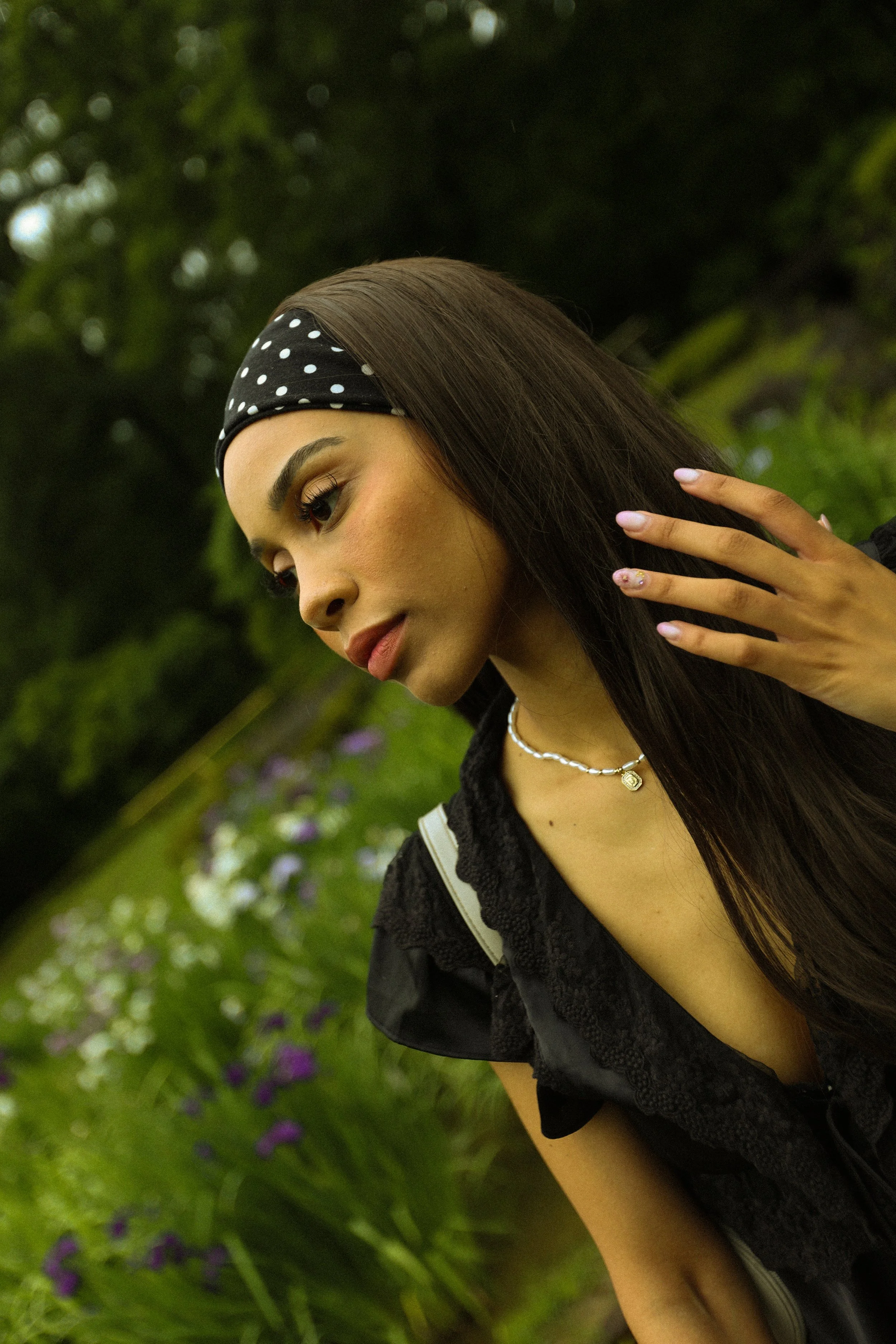 A young woman with long straight dark hair, wearing a black polka-dot headband, black top with lace detail, and a pearl necklace, sitting outdoors among green trees and purple flowers, touching her hair with her right hand.