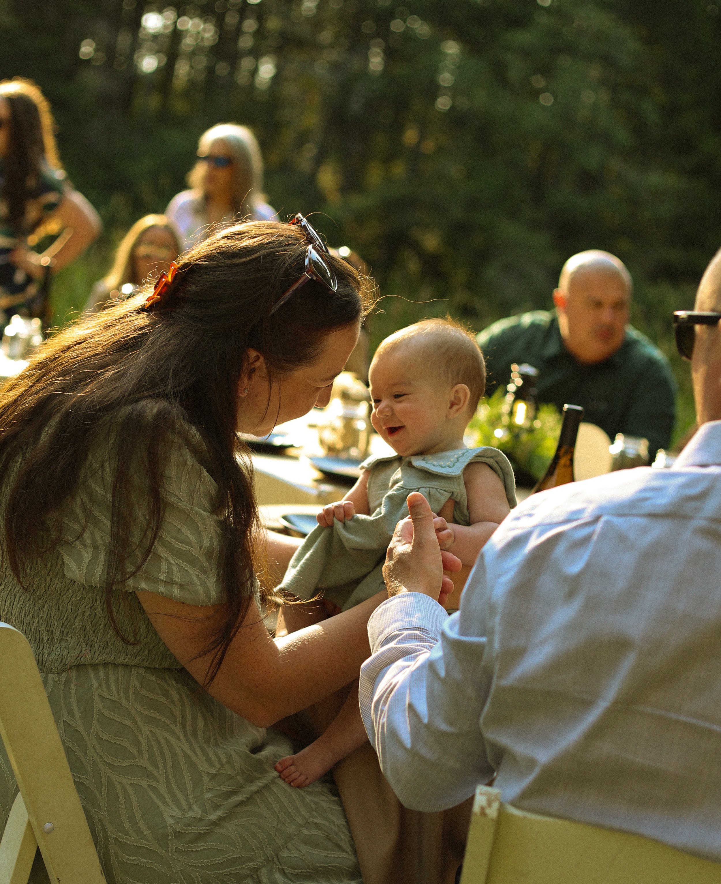 A woman with long dark hair, sunglasses on her head, and a patterned green dress is holding a smiling baby in her arms. They are outdoors, with other people blurred in the background and trees with sunlight filtering through the leaves.