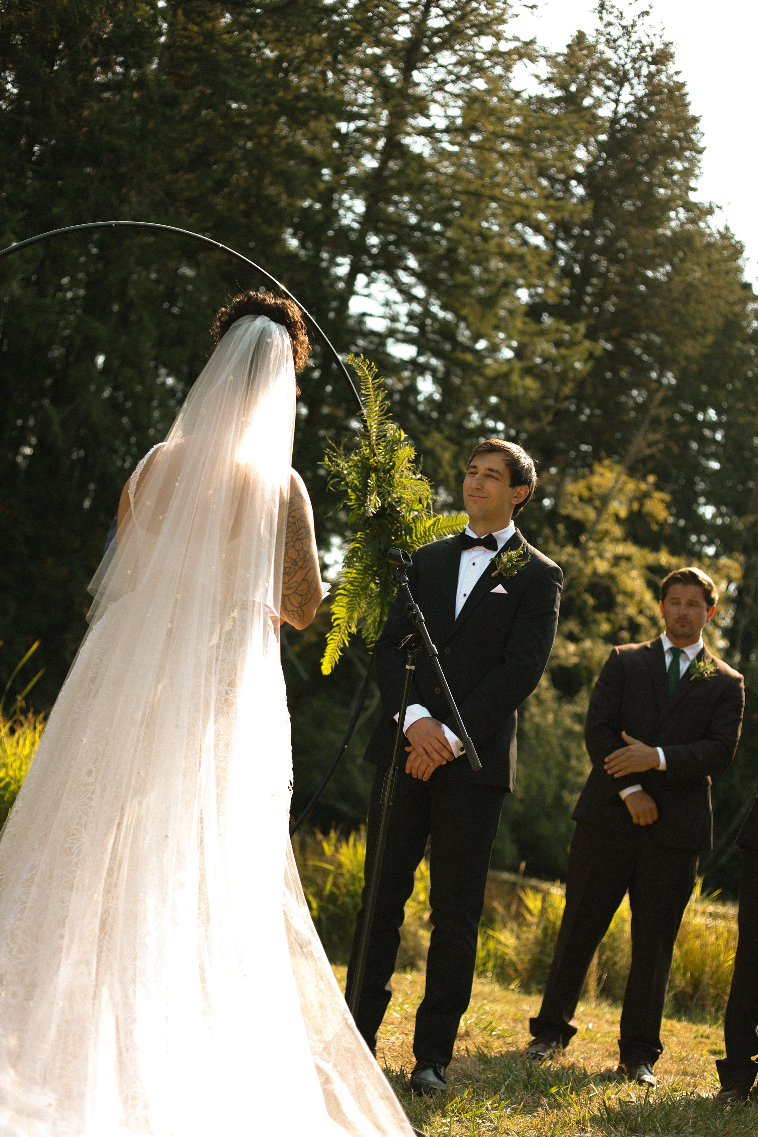 A bride with a veil and tattoo on her arm stands in front of a groom dressed in a tuxedo during an outdoor wedding ceremony, with other men in formal attire in the background, surrounded by trees and greenery.