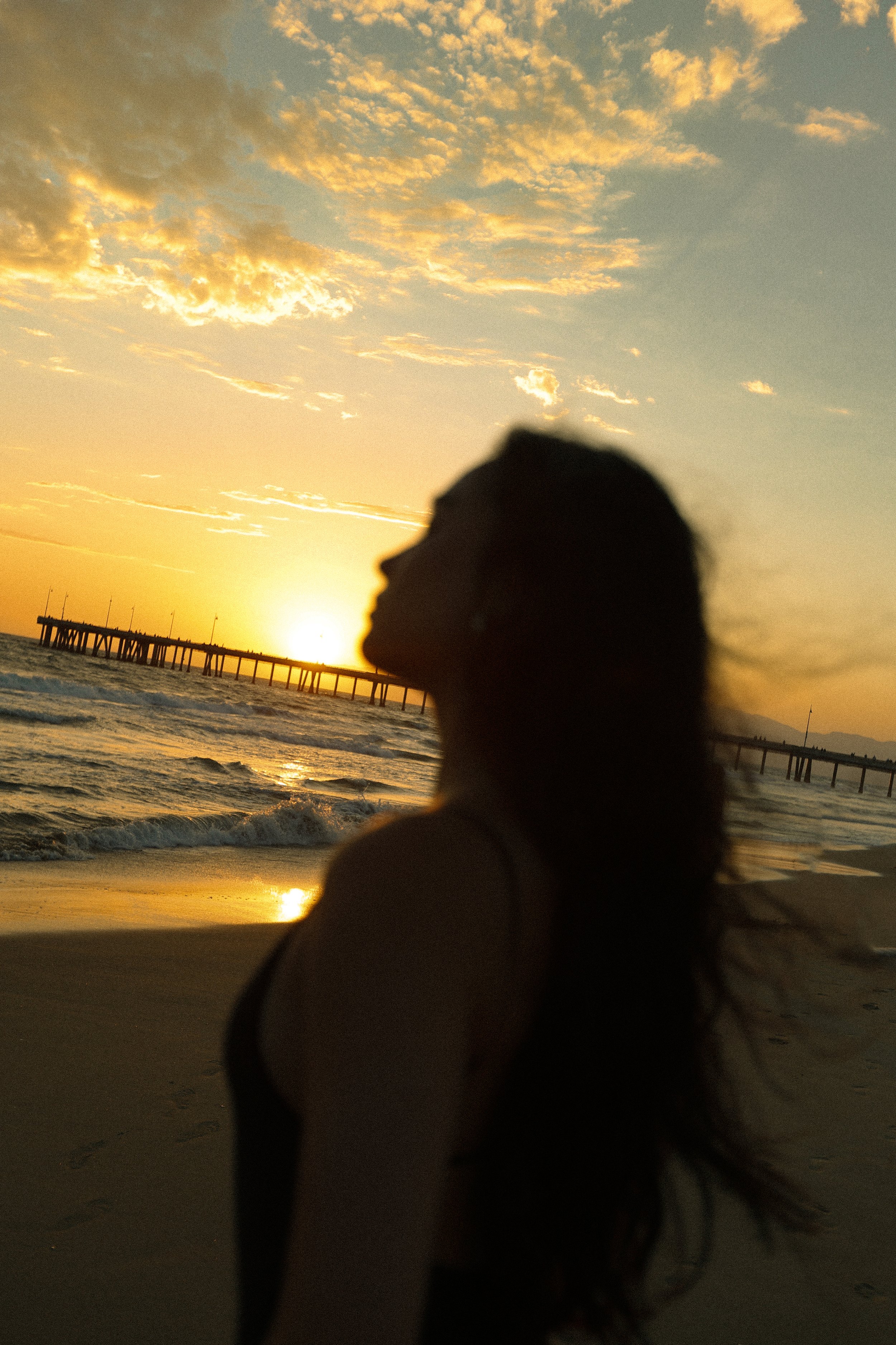 A person with long hair standing on the beach at sunset with clouds in the sky and a pier in the background.