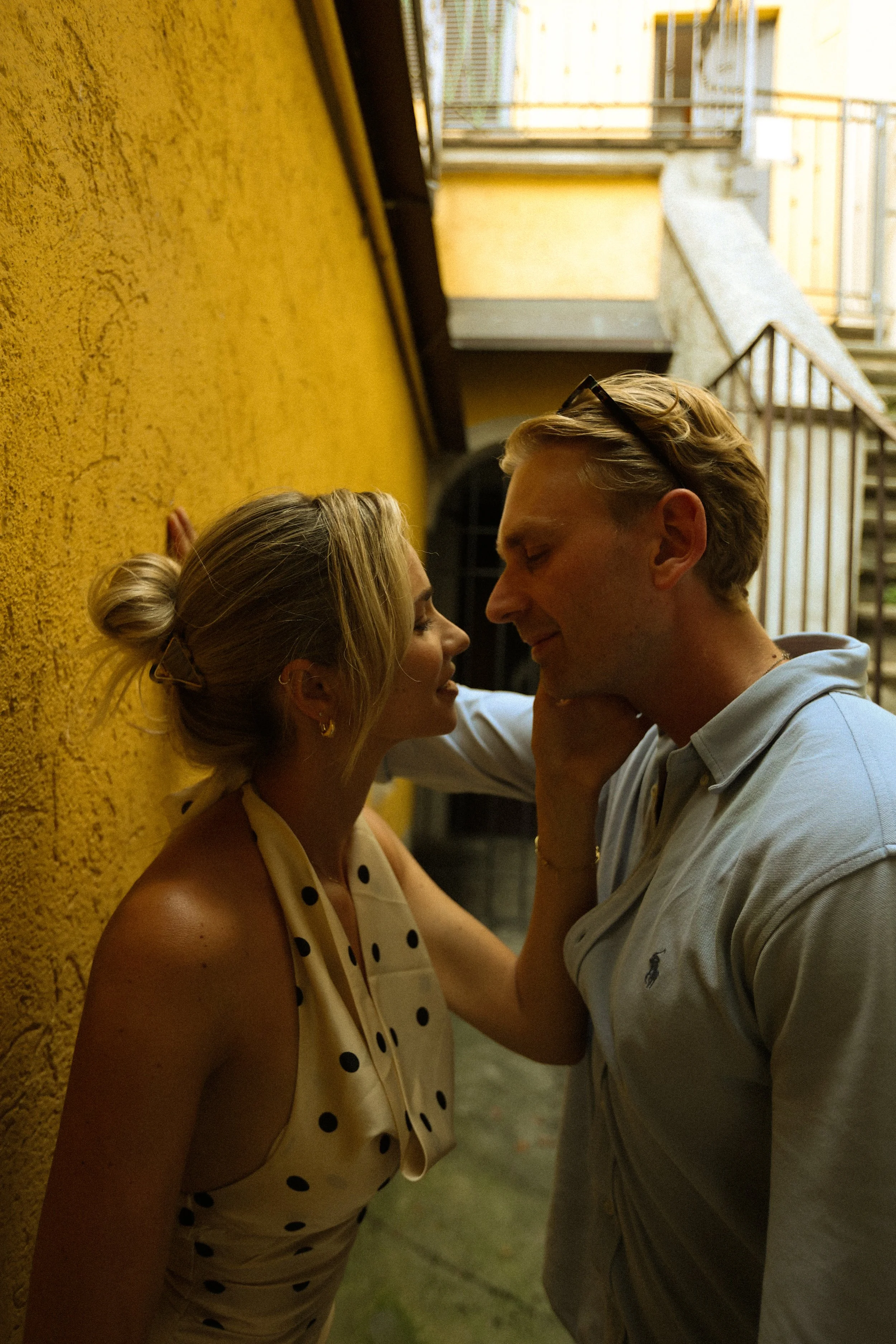 A man and a woman leaning close to each other against a yellow wall, smiling and looking into each other's eyes. The woman has blonde hair tied in a bun and is wearing a polka dot dress; the man has blond hair and is wearing glasses and a light-color