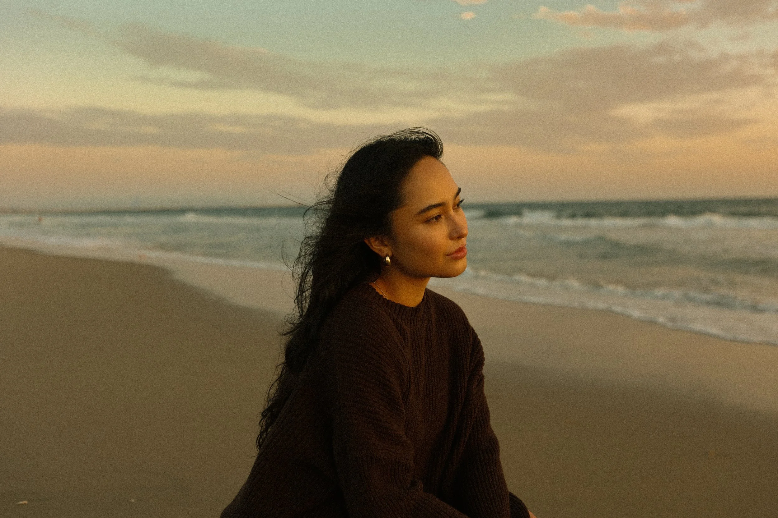 A woman with dark hair and earrings sitting on the beach facing the ocean during sunset or sunrise.