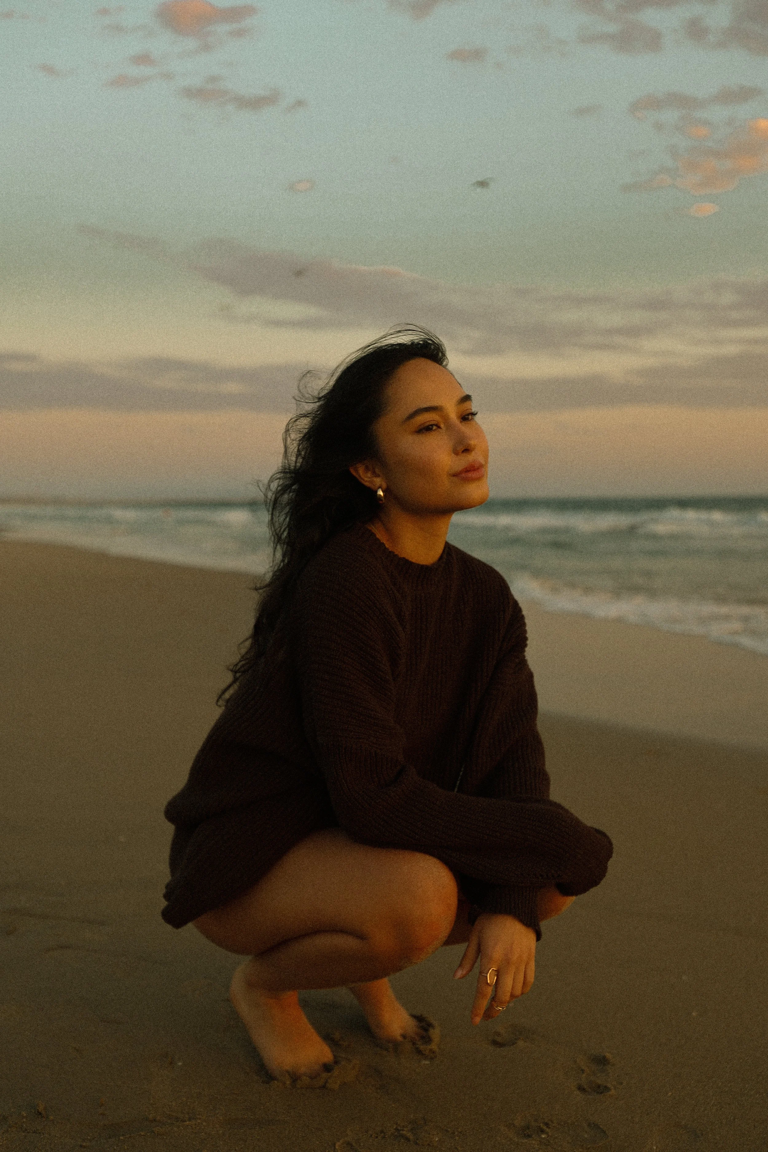 A woman crouching on the beach with her hair blowing in the wind during sunset, wearing a dark sweater and rings on her fingers.