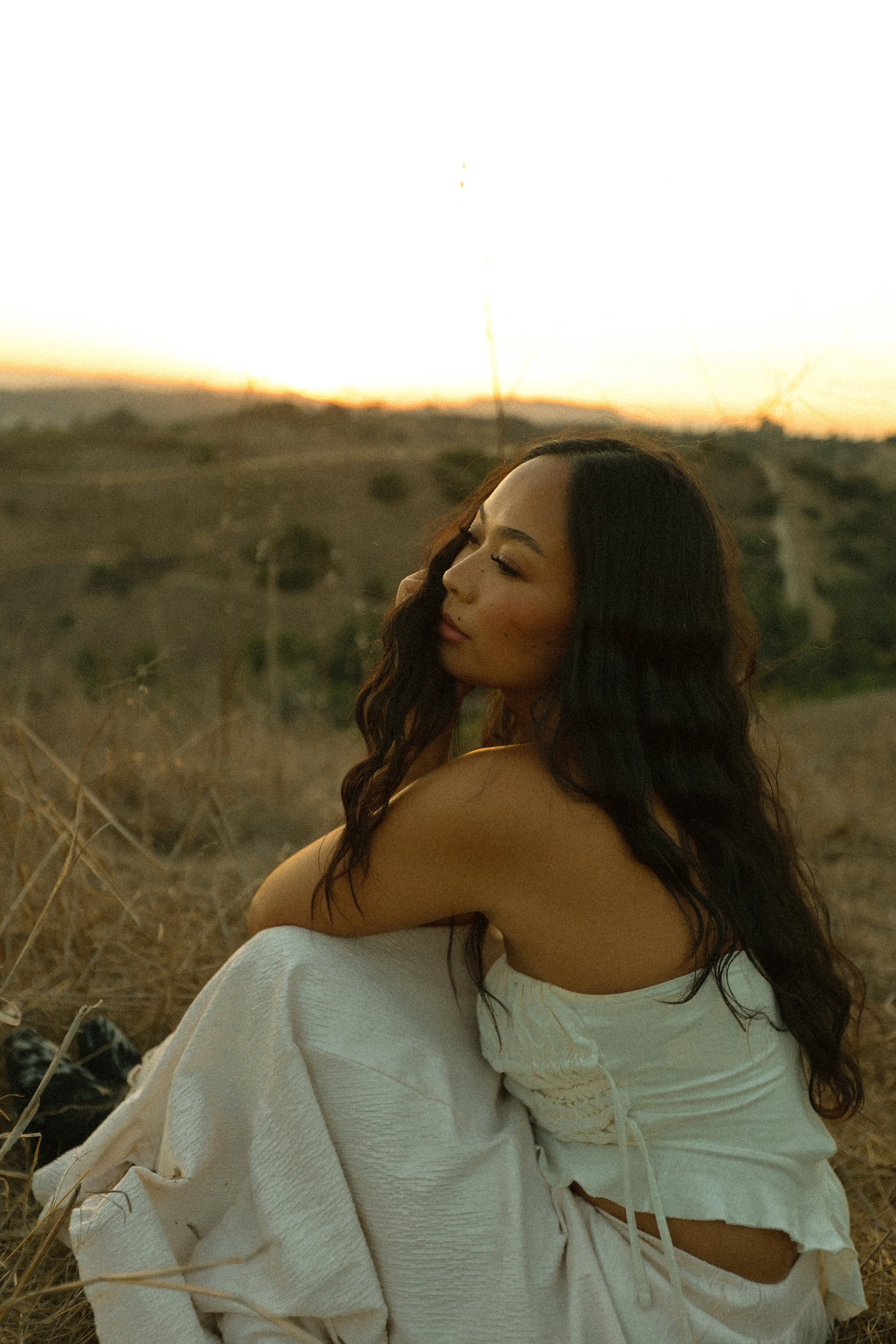 A woman sitting outdoors during sunset, with a dry landscape in the background, wearing a white outfit and with long dark hair.