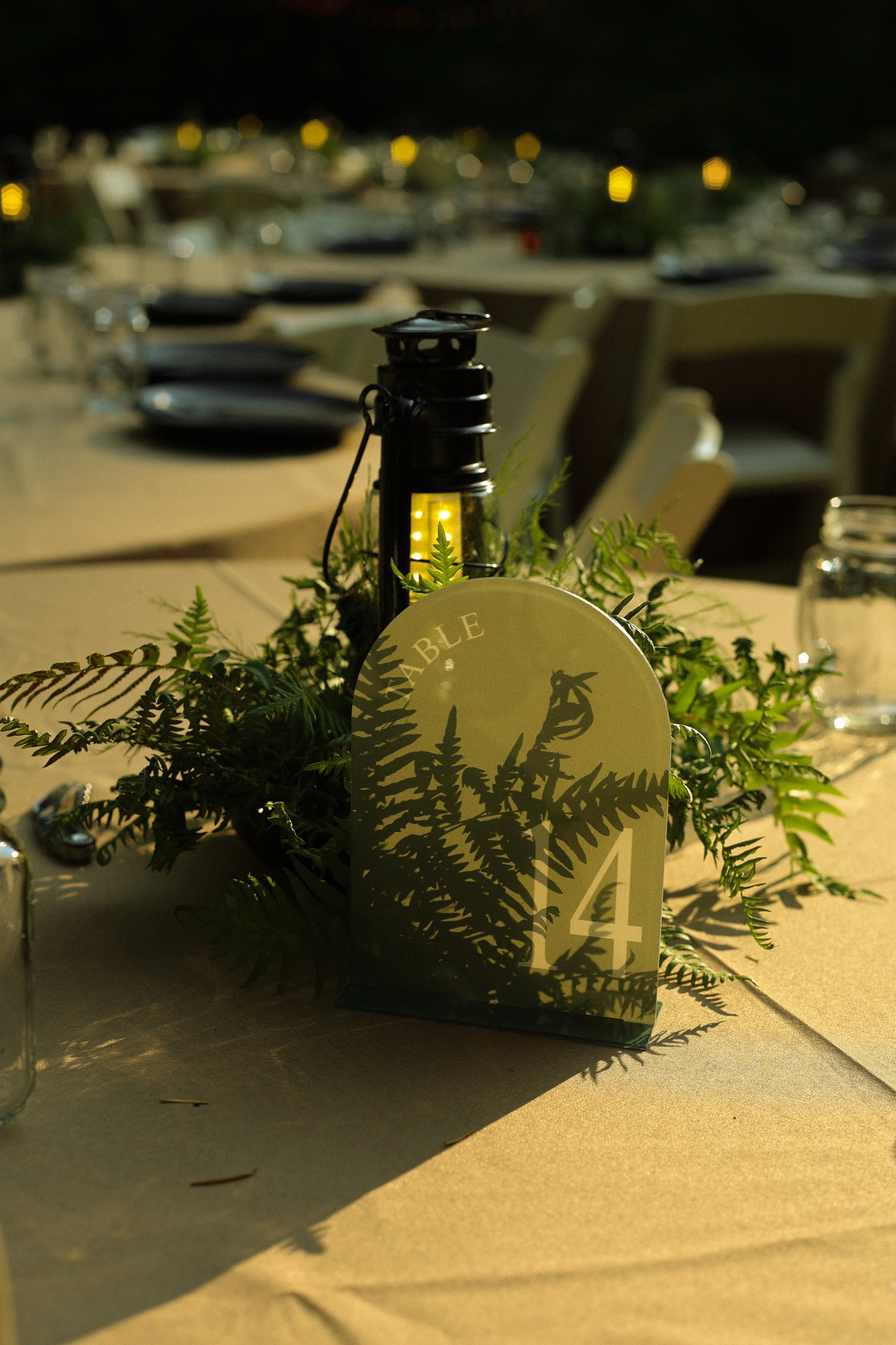 A close-up of a table centerpiece with a small lantern light, surrounded by greenery, set on a table with a table number visible. In the background, blurred tables and chairs are visible at night.
