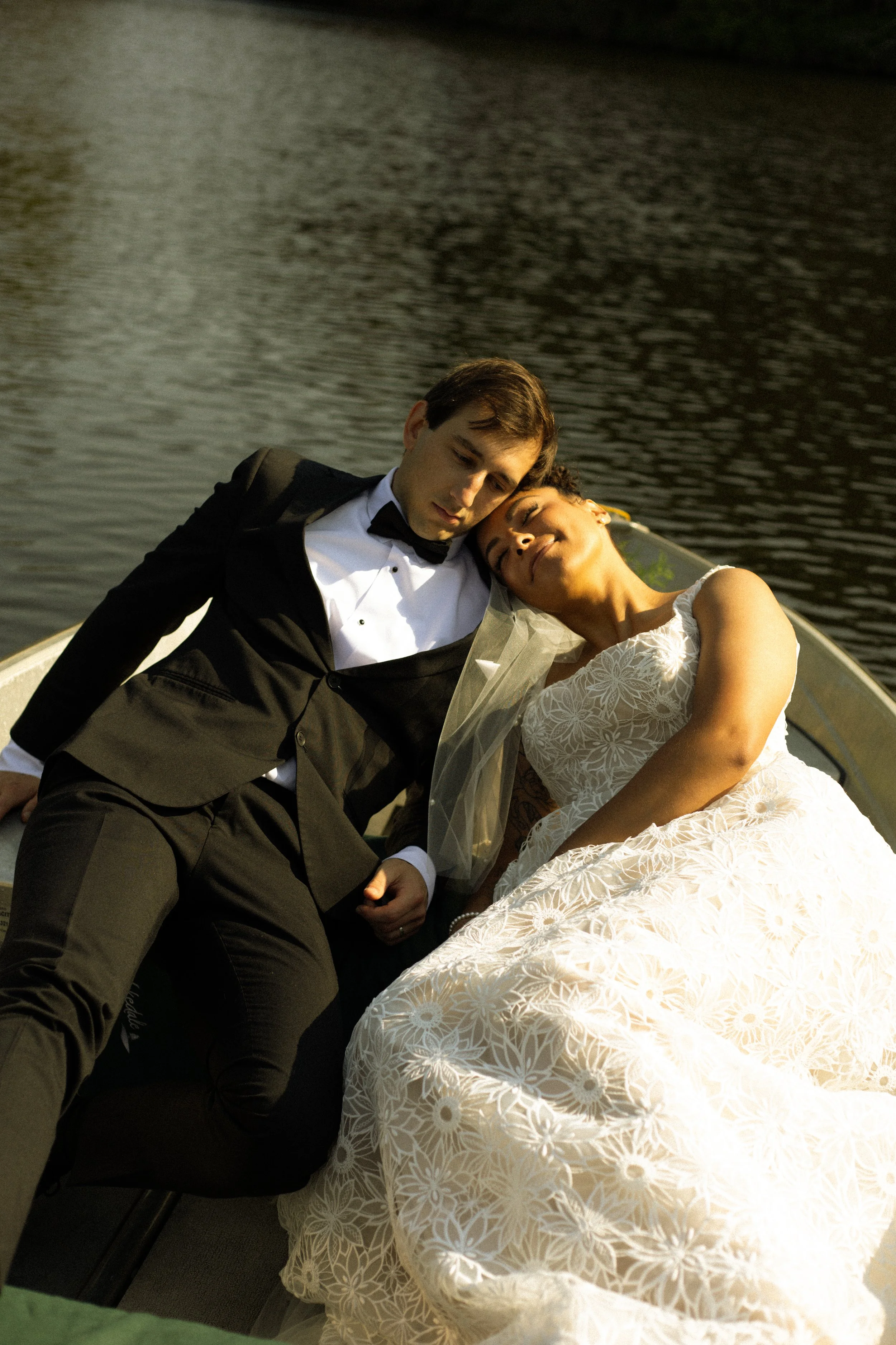 A man in a tuxedo and a woman in a white lace dress are on a boat near a body of water, resting their heads together with their eyes closed.