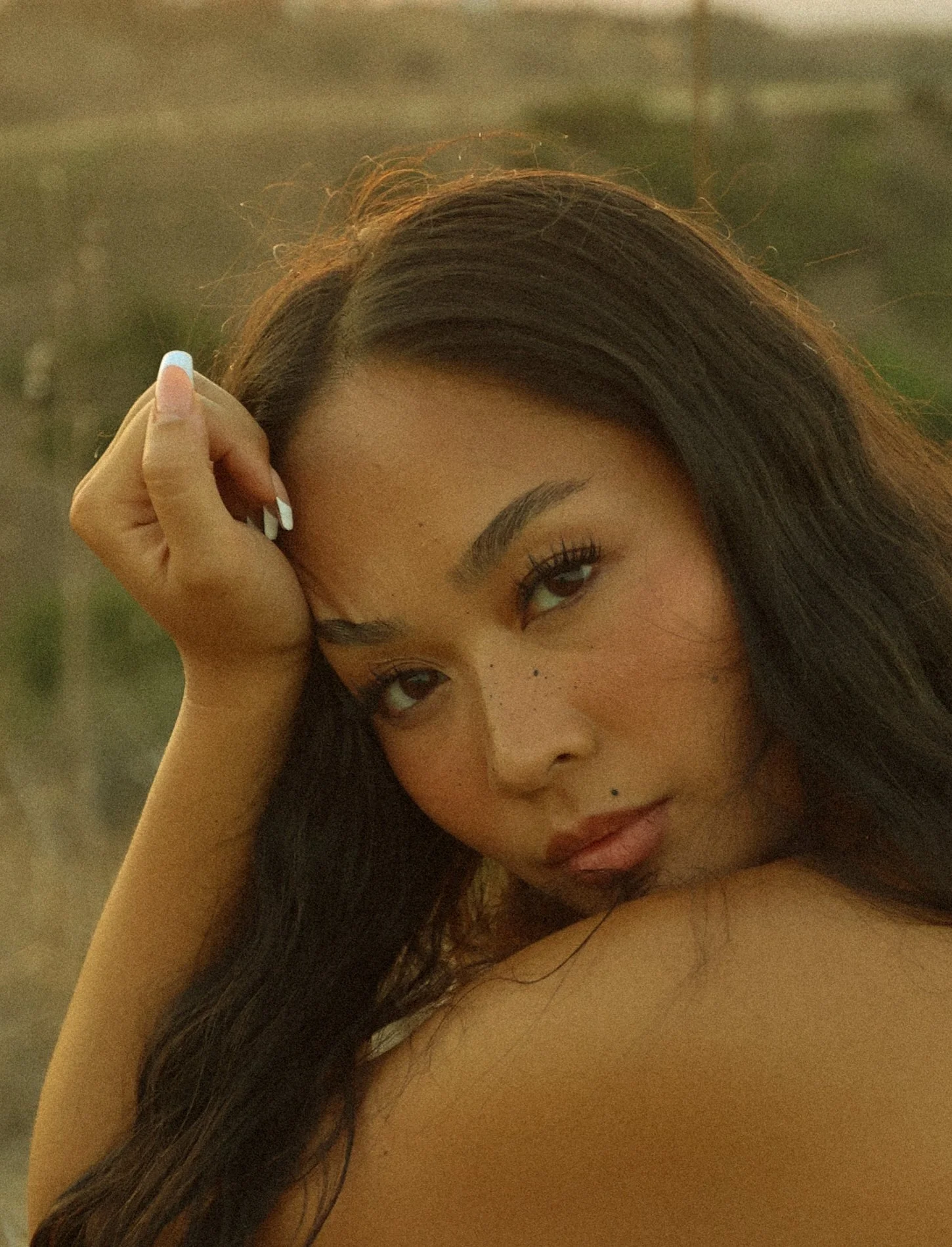 A woman with long dark hair resting her hand on her forehead, looking at the camera with a soft expression.