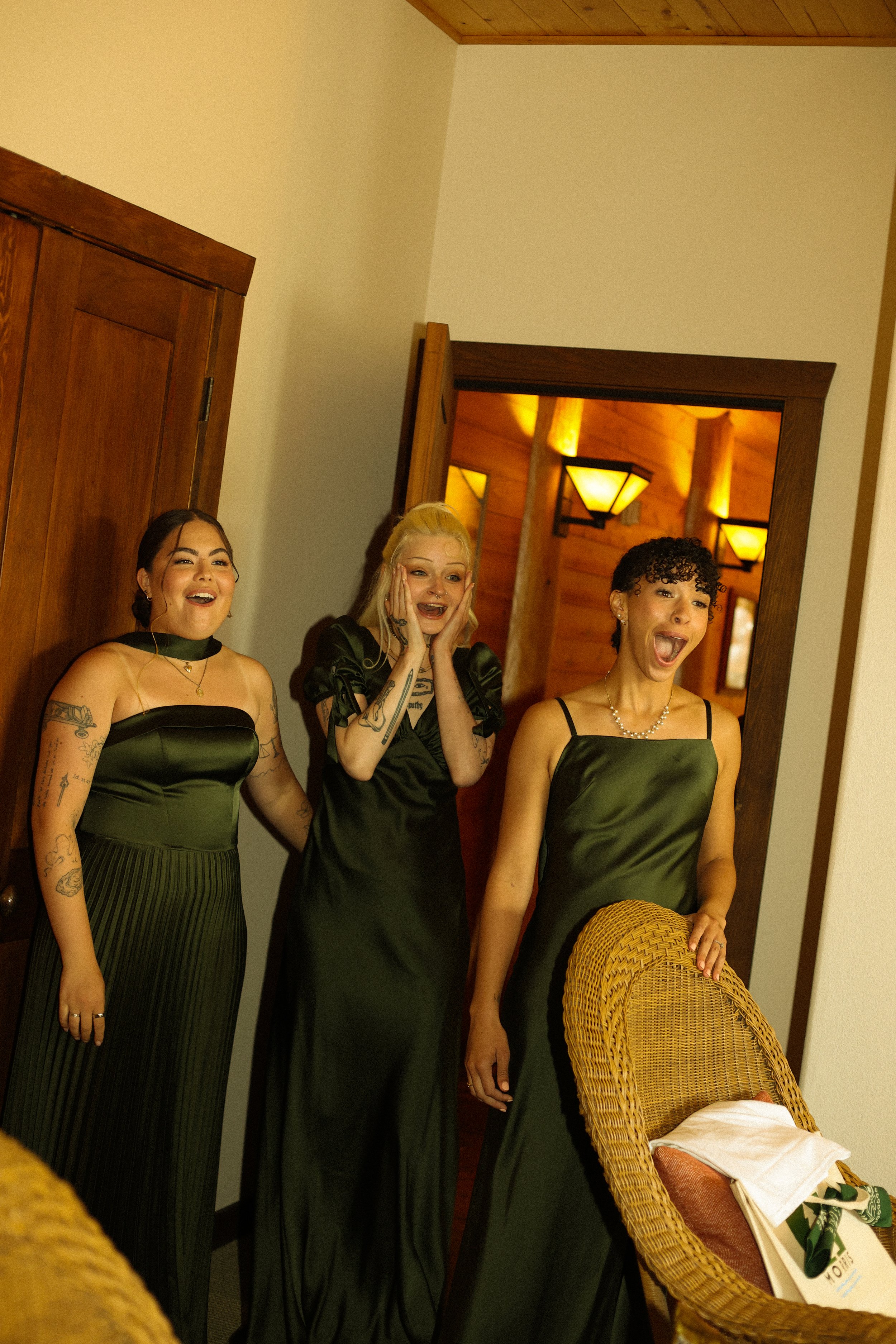 Four women in black dresses standing in front of a mirror with wooden trim, some showing surprised or excited expressions. The setting appears to be indoors with warm lighting and a wooden ceiling.