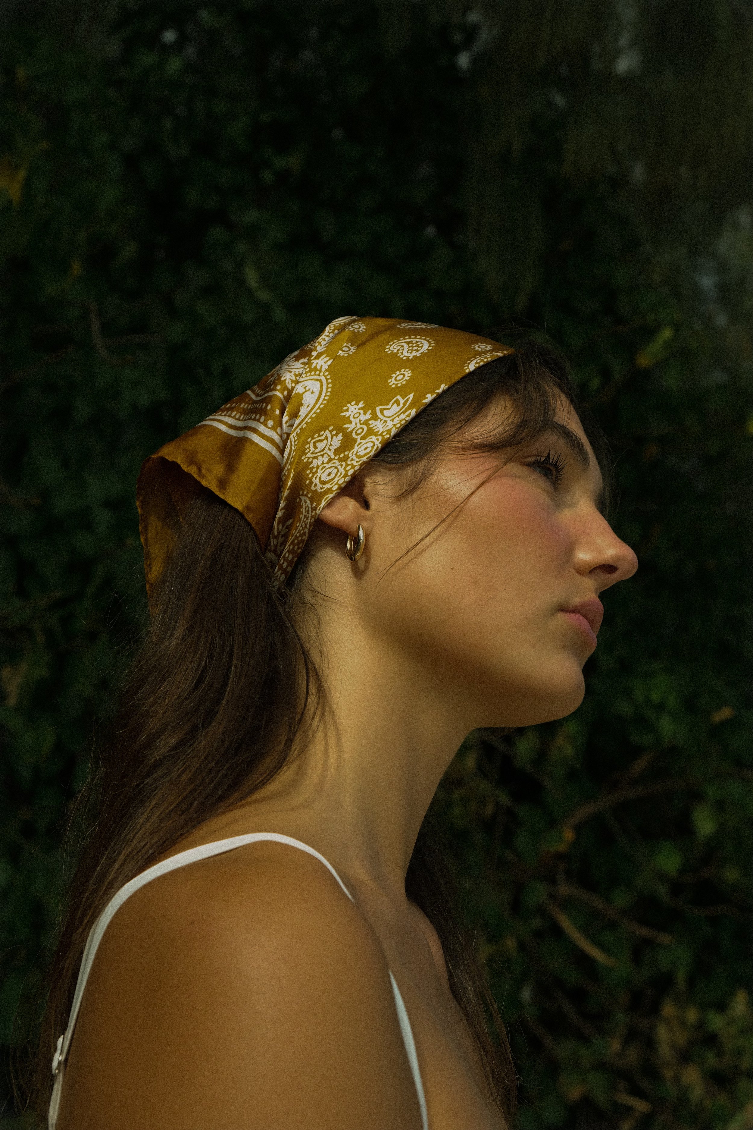 A woman with long brown hair wearing a brown and white patterned headscarf, earrings, and a white tank top, standing outdoors in front of greenery.