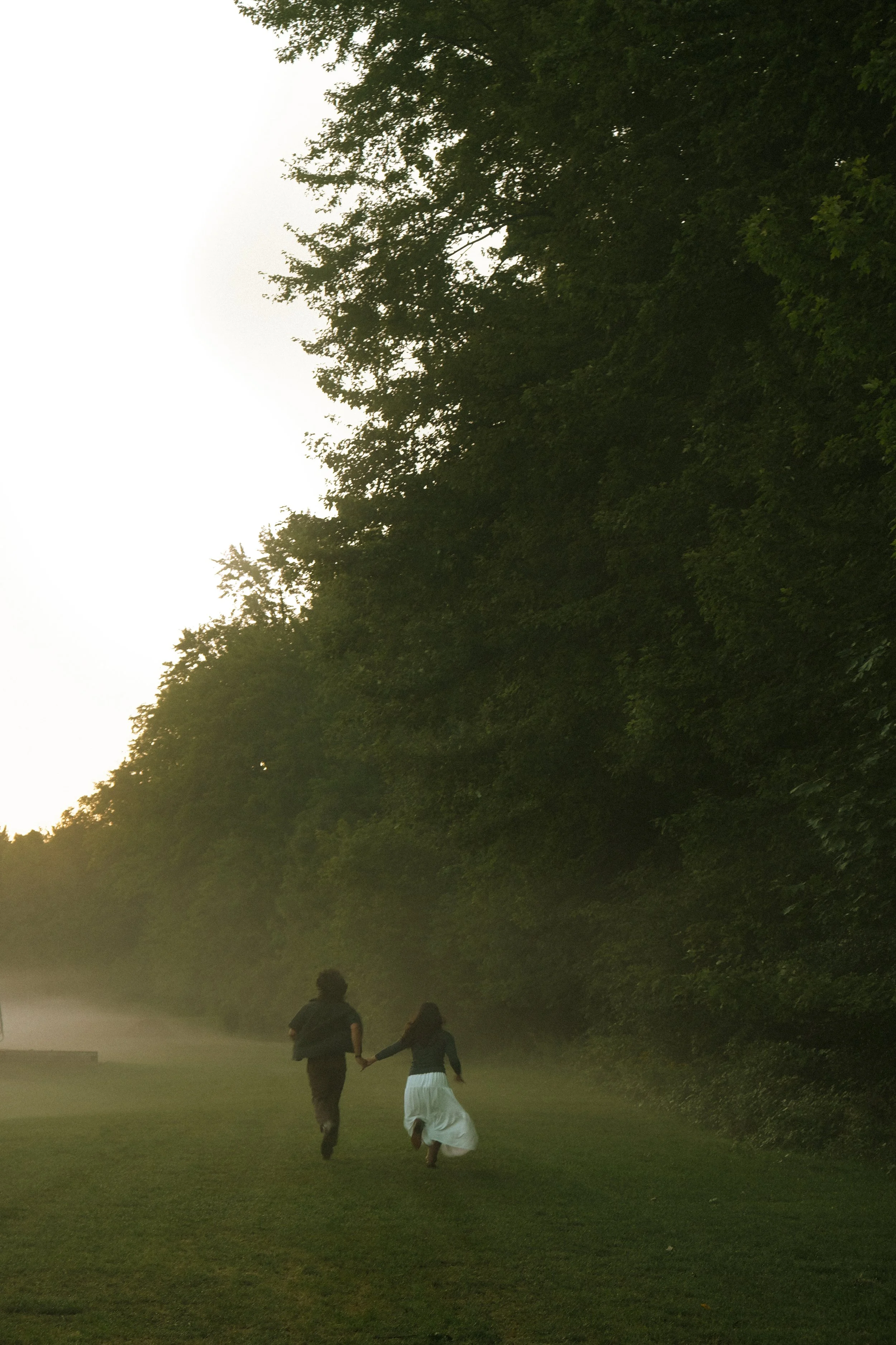 A couple holding hands and walking along a grassy path in a park during sunset, surrounded by dense trees with green foliage.