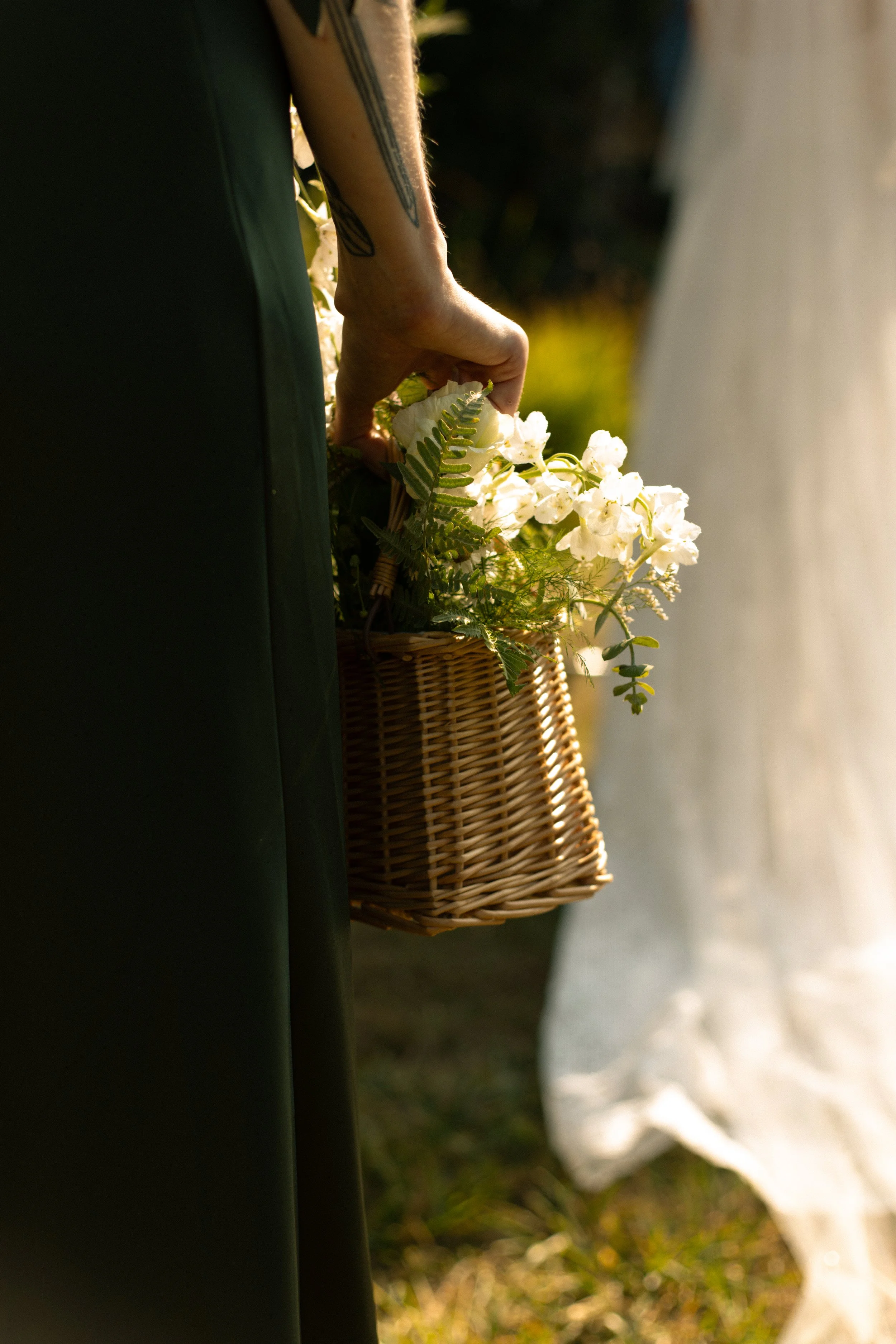 A hand reaching into a wicker basket filled with white flowers and greenery, with a dark background and a white cloth draped nearby.