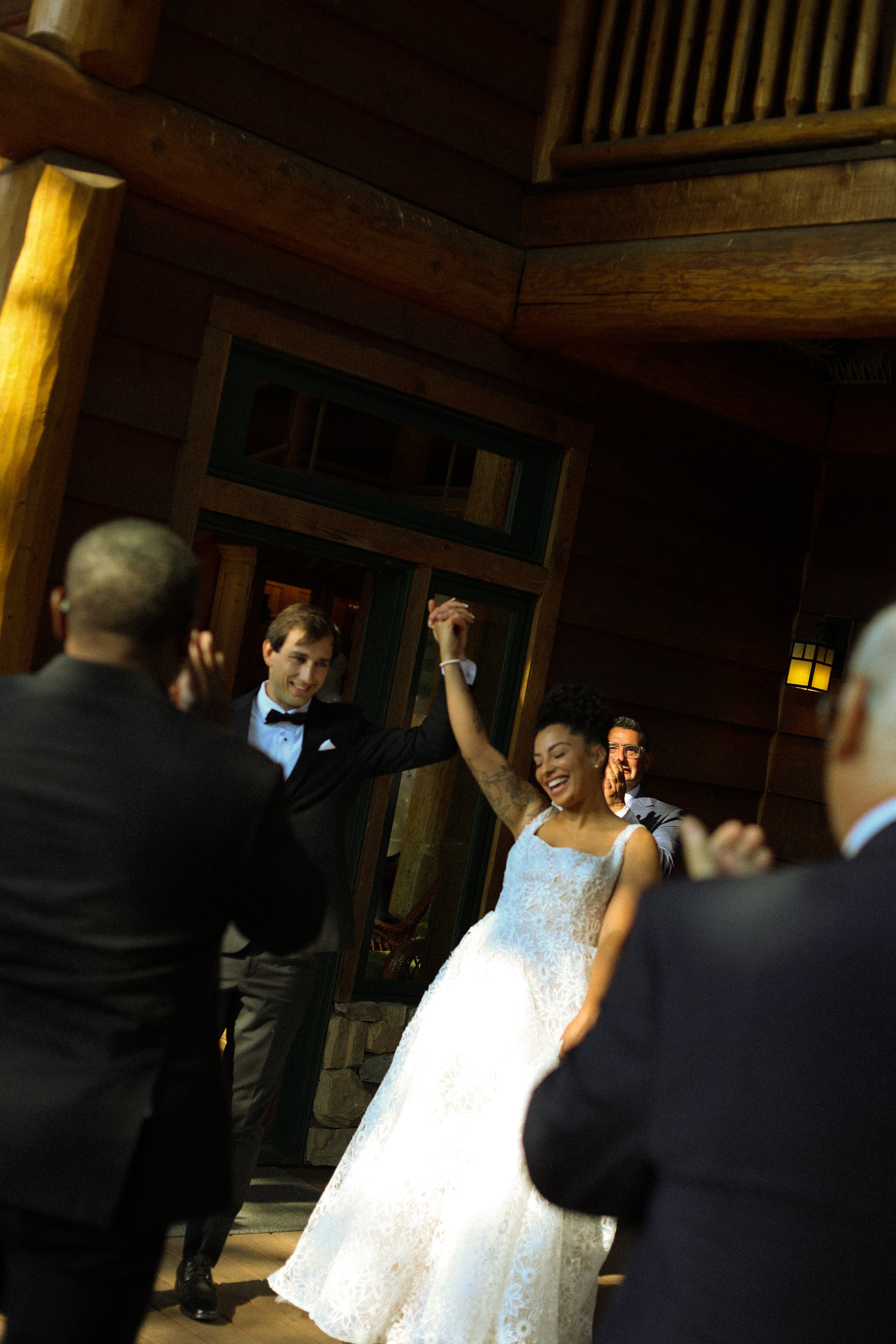 A wedding celebration with a bride in a white lace gown and five men in tuxedos dancing and smiling in a warmly lit wooden venue.