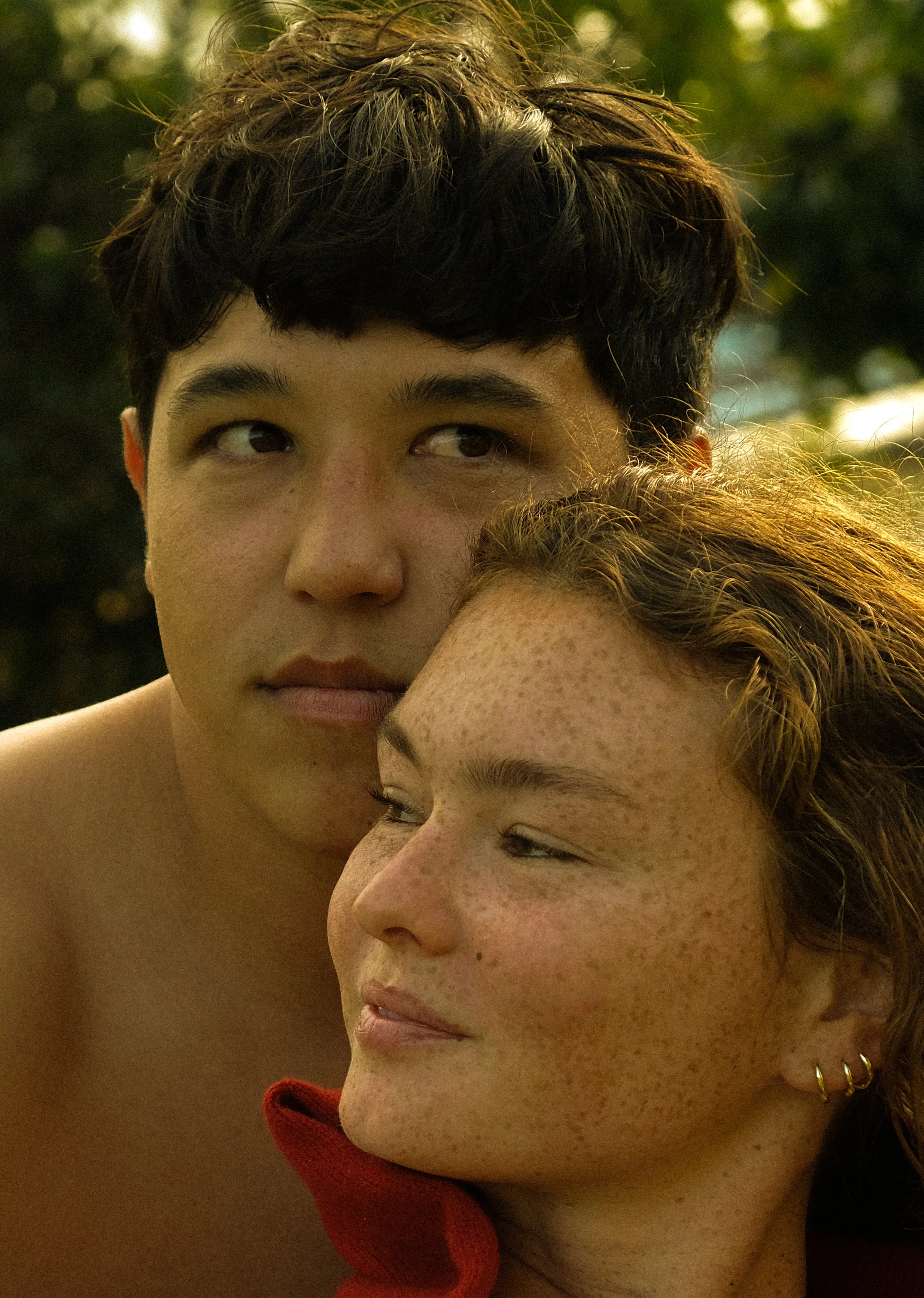 Close-up of two young women with their faces pressed close together, one with short dark hair and the other with curly red hair, both with relaxed expressions against a blurred outdoor background with sunlight.