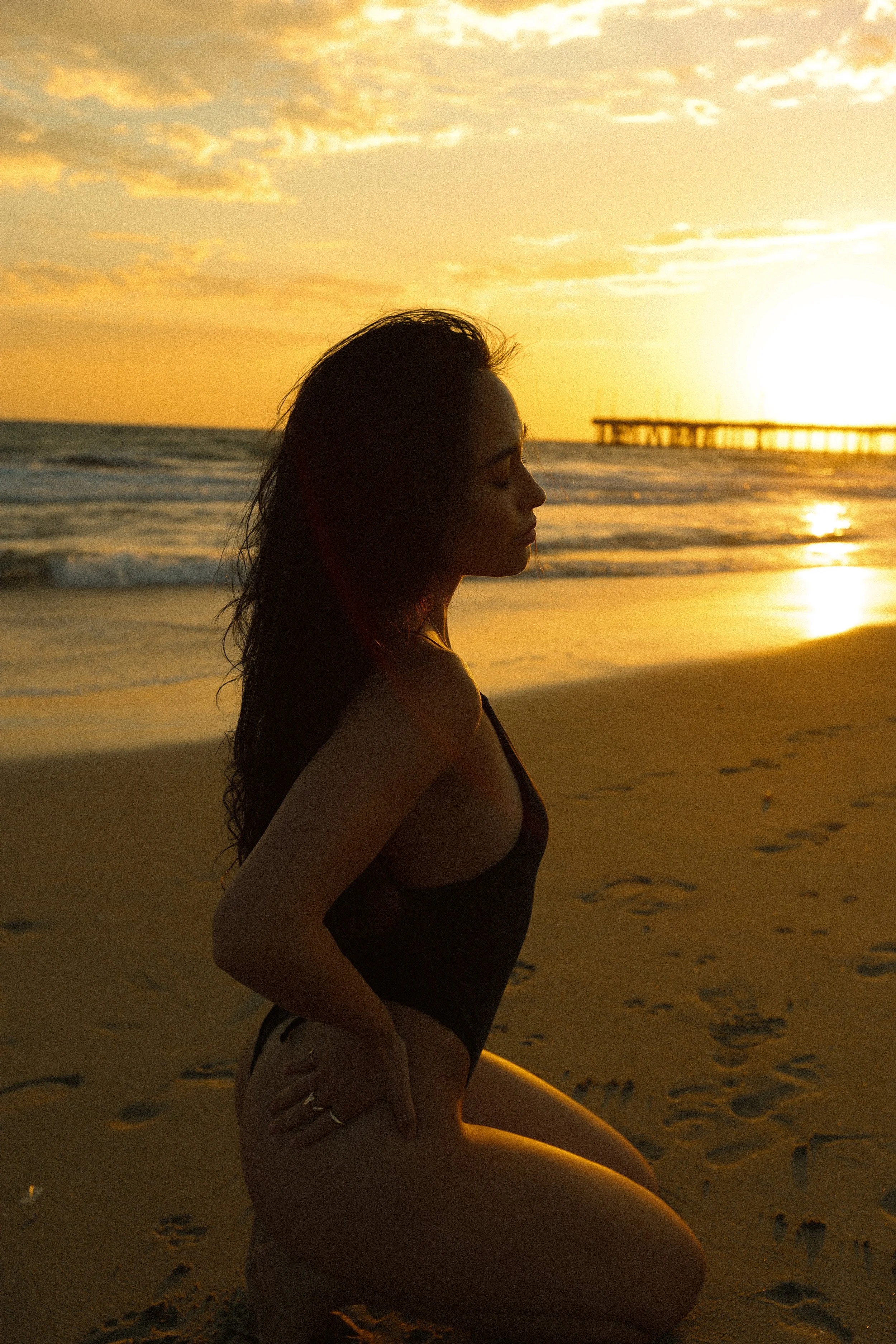 A woman with long hair in a one-piece swimsuit kneeling on the sand at the beach during sunset, with a pier in the background and the sun near the horizon.