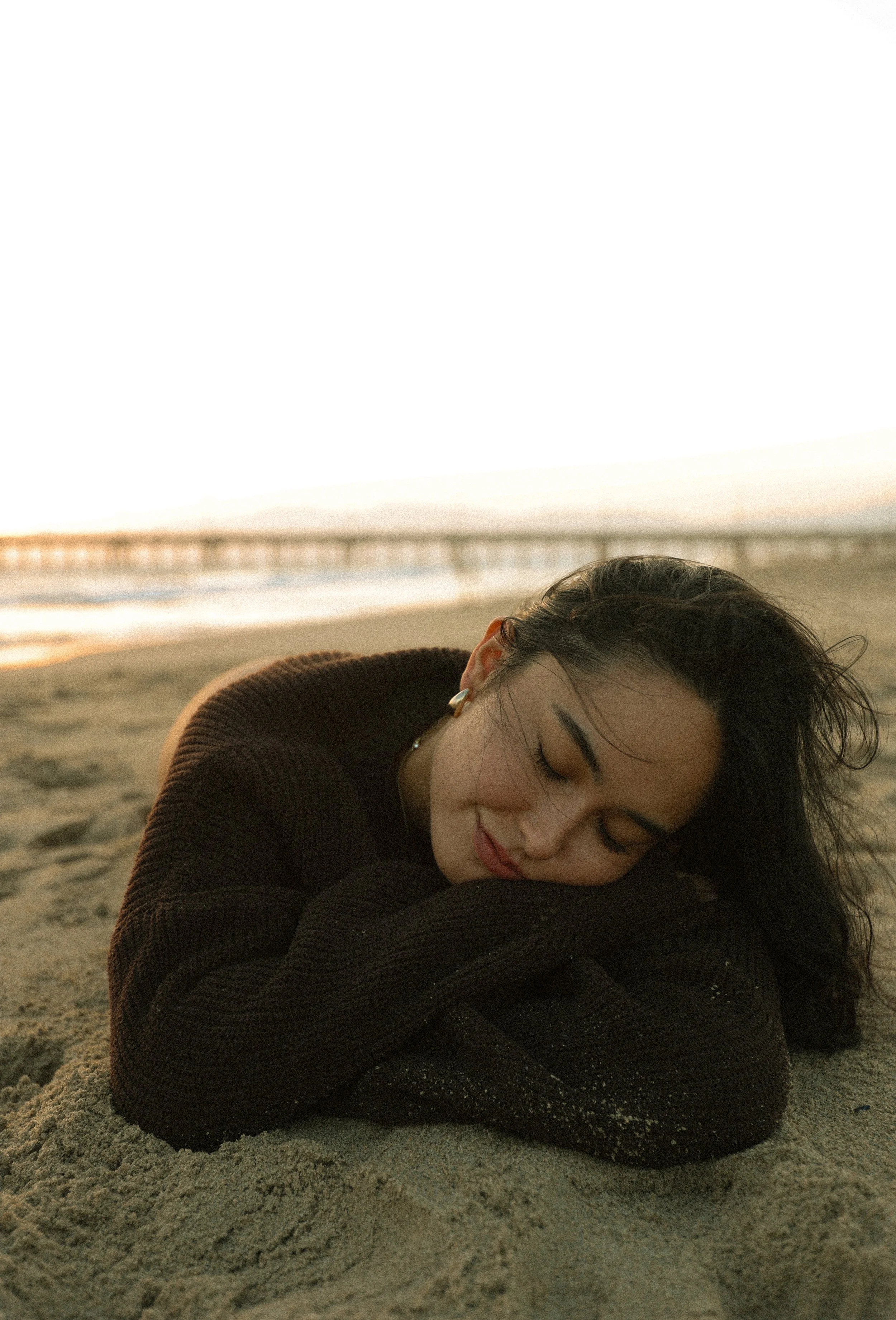 A woman with dark hair and earrings rests her head on her arm on the sandy beach during sunset or sunrise, with a pier in the background.