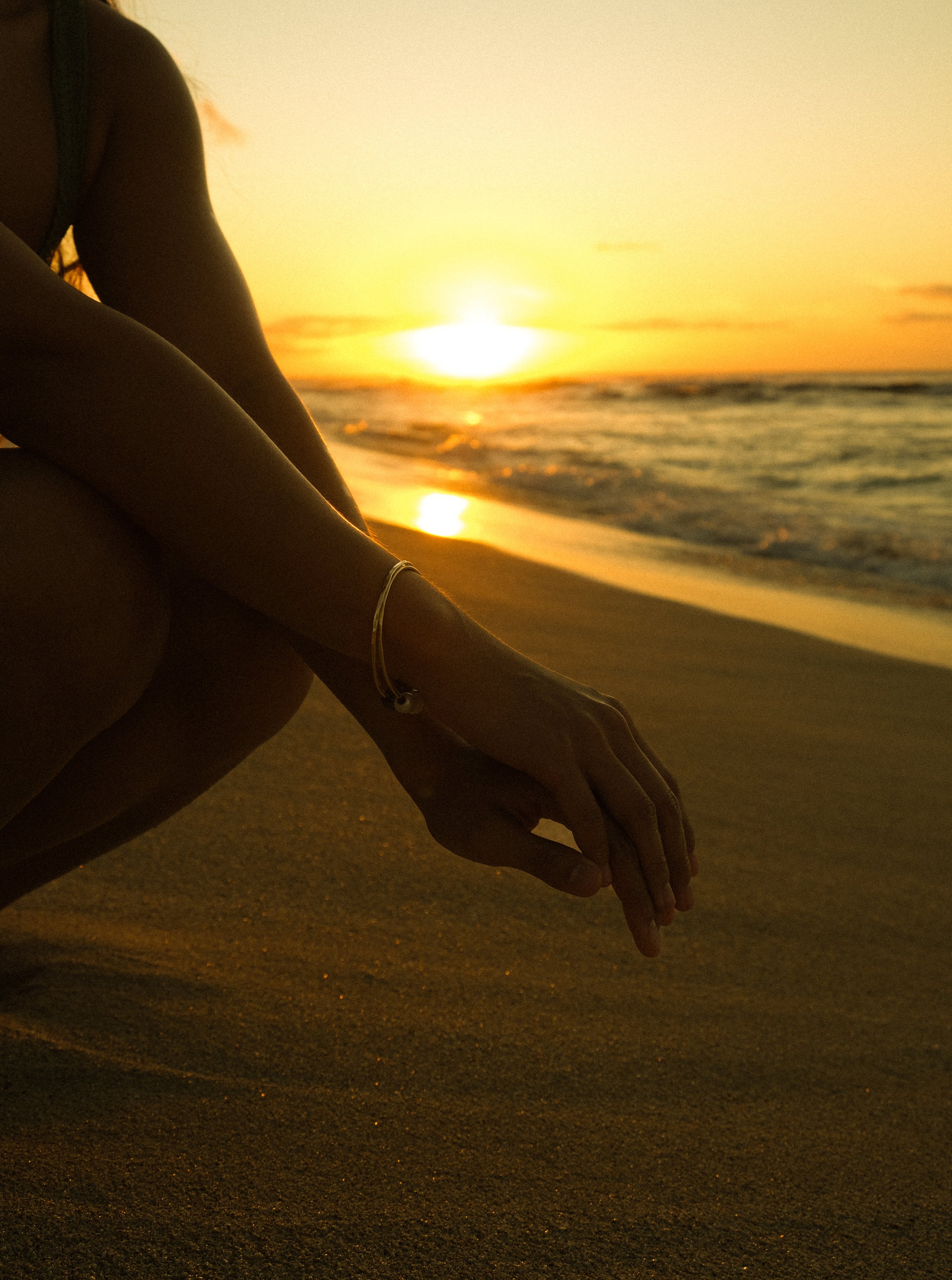 Silhouette of a person sitting on the beach during sunset, with their hand resting on their knee and wearing a bracelet, in front of the ocean with the sun setting on the horizon.