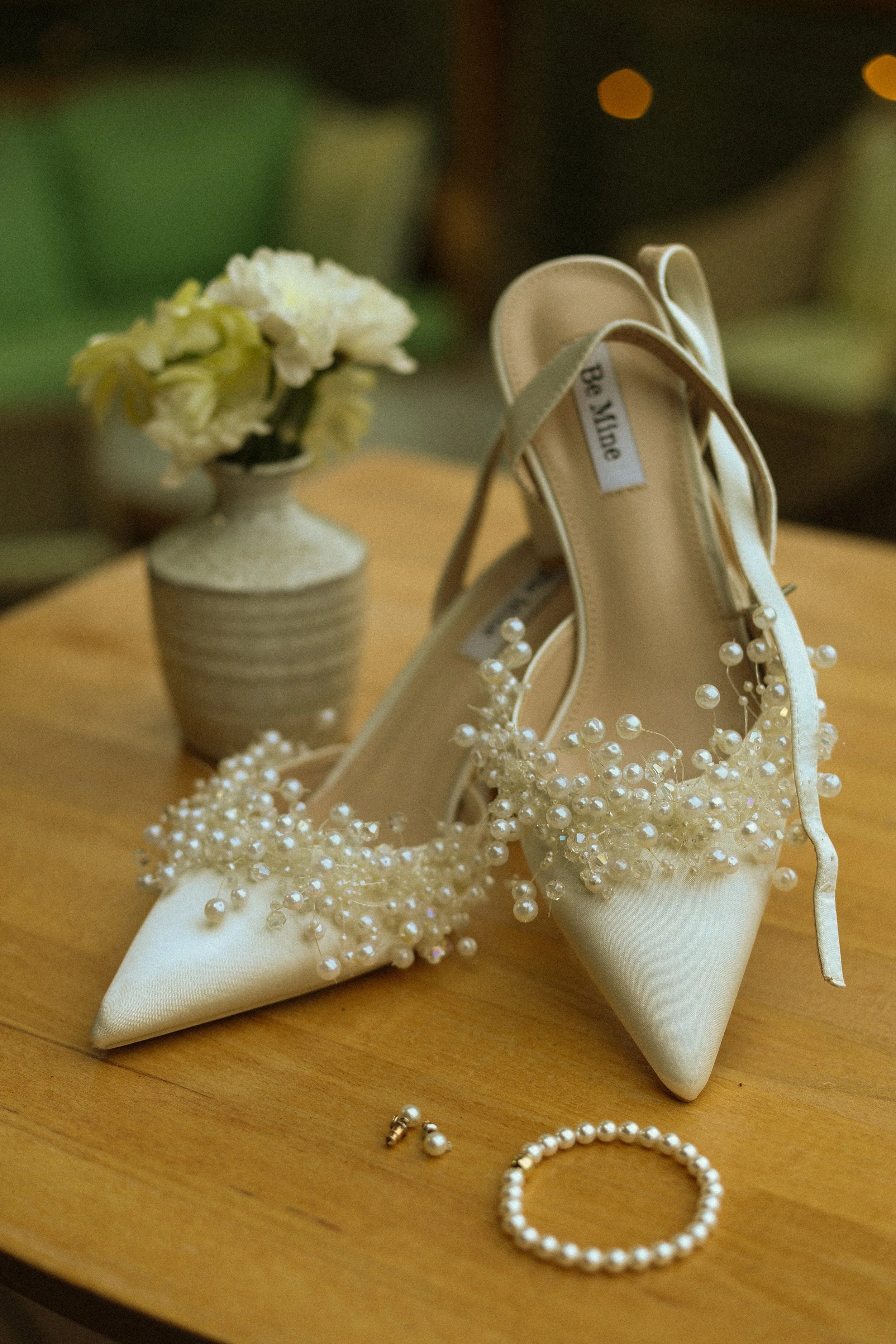 A pair of white satin bridal shoes decorated with pearls, placed on a wooden surface. Next to the shoes is a pearl bracelet and earrings. In the background, there is a small flower arrangement in a white textured vase, with a green cloth and warm out