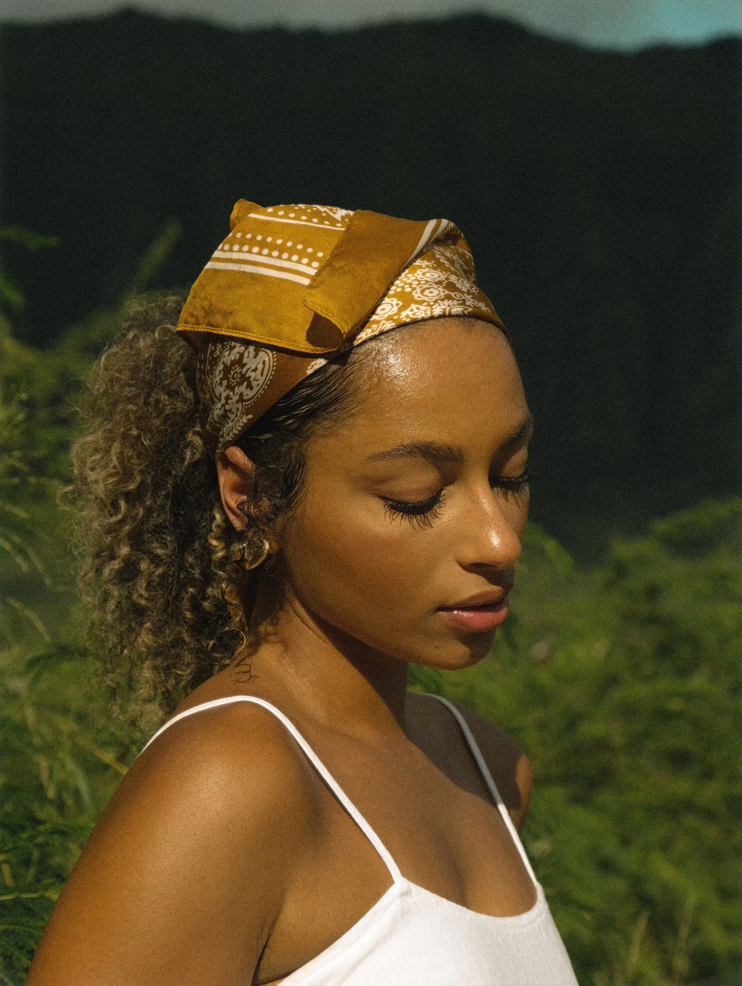 Close-up of a woman with curly hair, wearing a yellow and white patterned headscarf and a white spaghetti strap top, outdoors at night.
