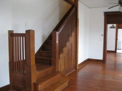 Wooden staircase with a banister in a room with hardwood flooring and white walls.